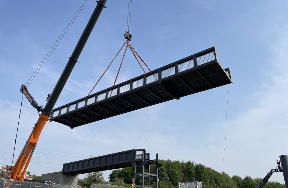 Crane lifting a large steel pedestrian bridge section at a construction site outdoors.