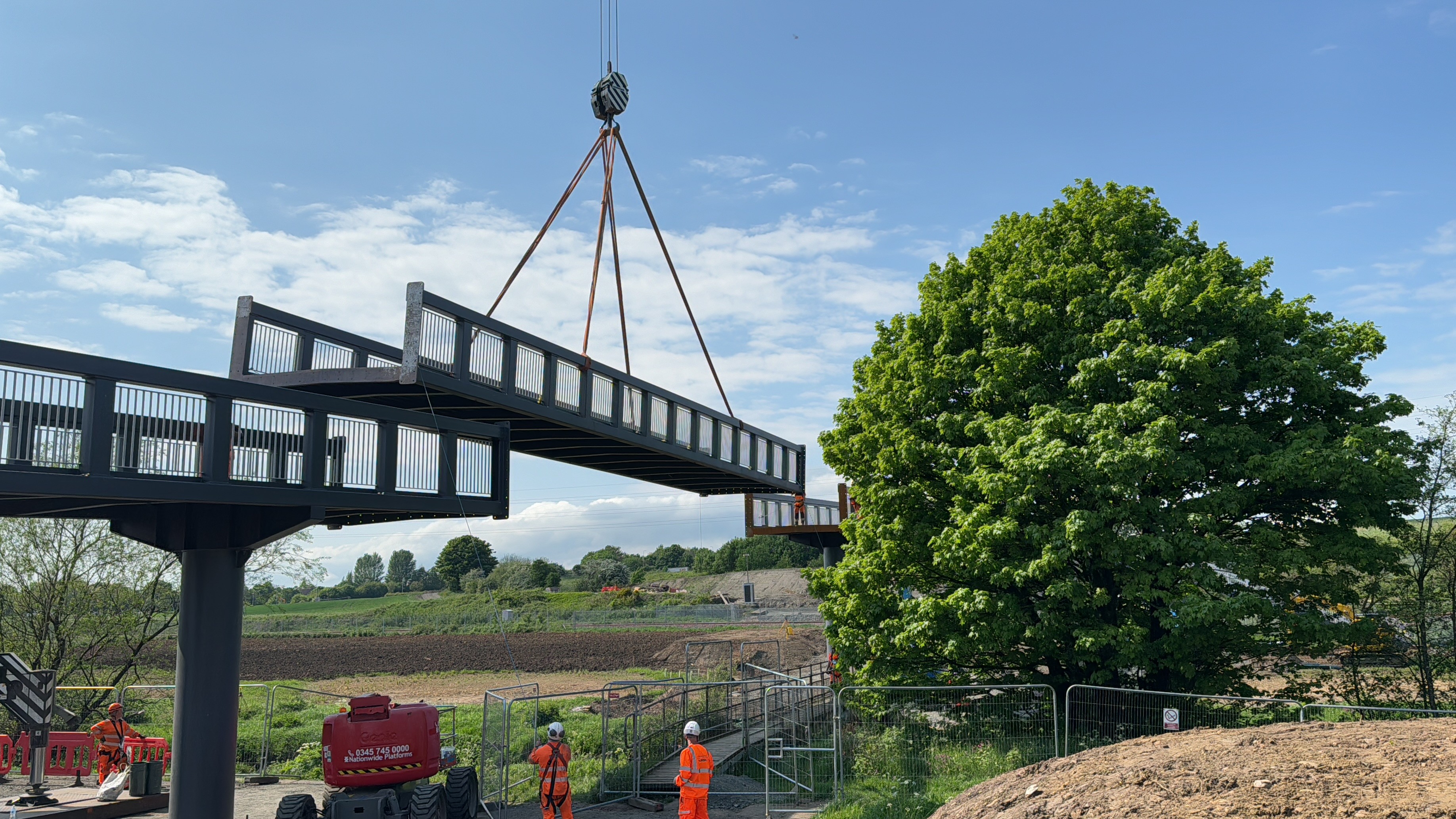 Construction workers installing a pedestrian bridge on a sunny day Crane lifting a metal bridge section at a construction site with workers and greenery.