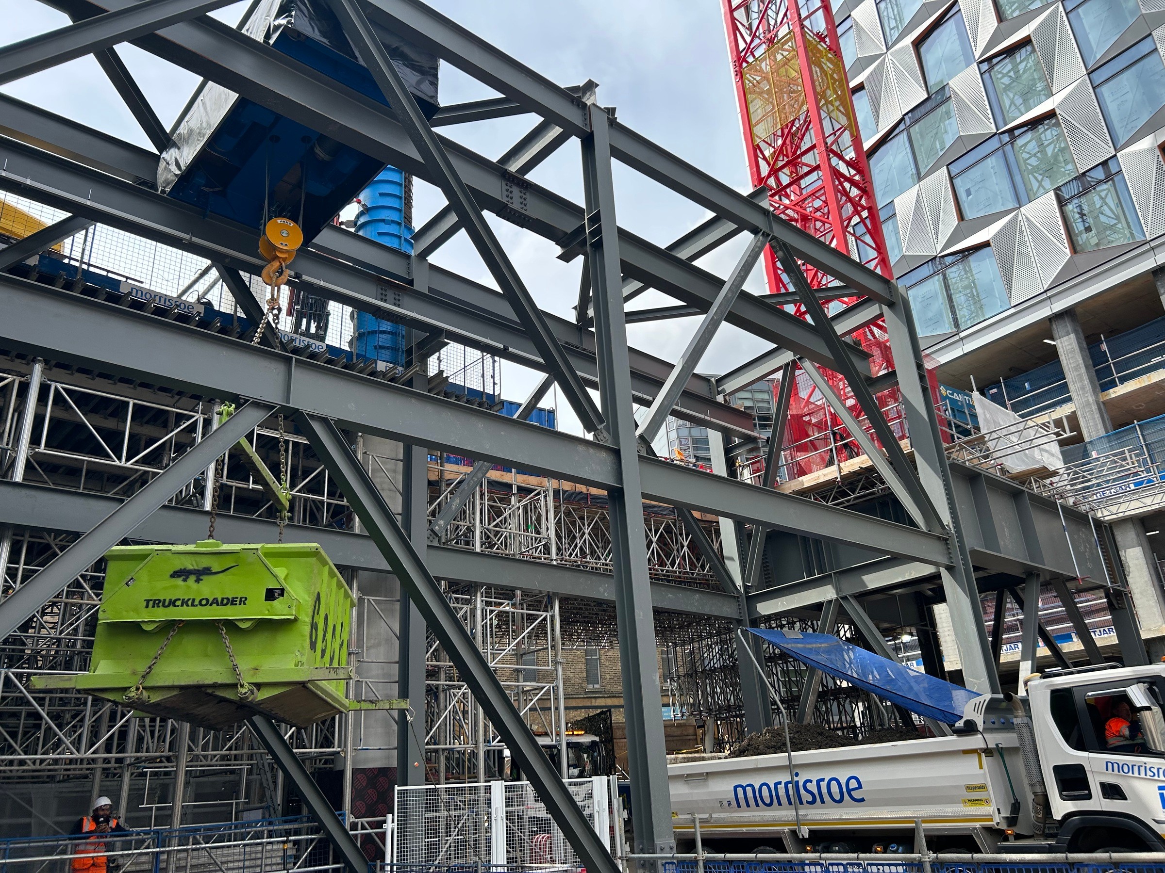 Construction site with steel framework, crane, and a truck loader in front of modern buildings.