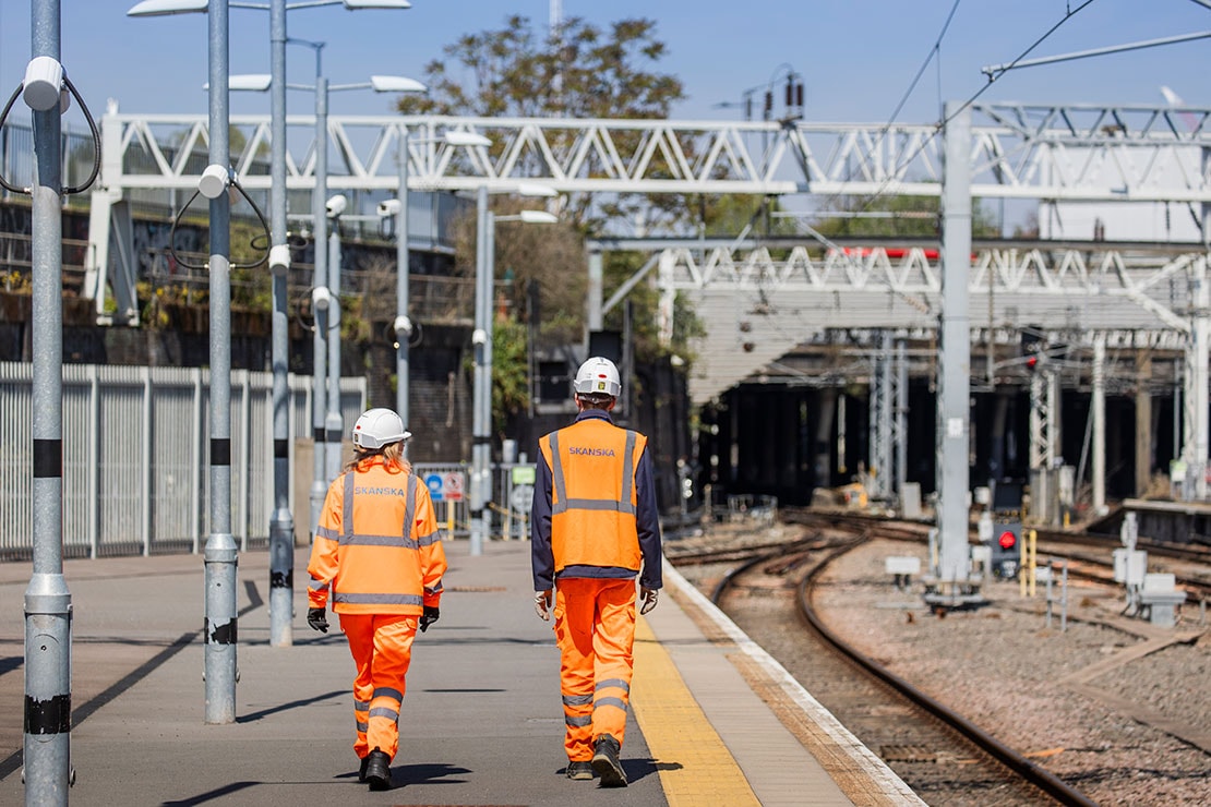 Railway Workers in Safety Gear Walking Along Train Platform Tracks Two railway workers in orange safety gear walking along a train station platform.