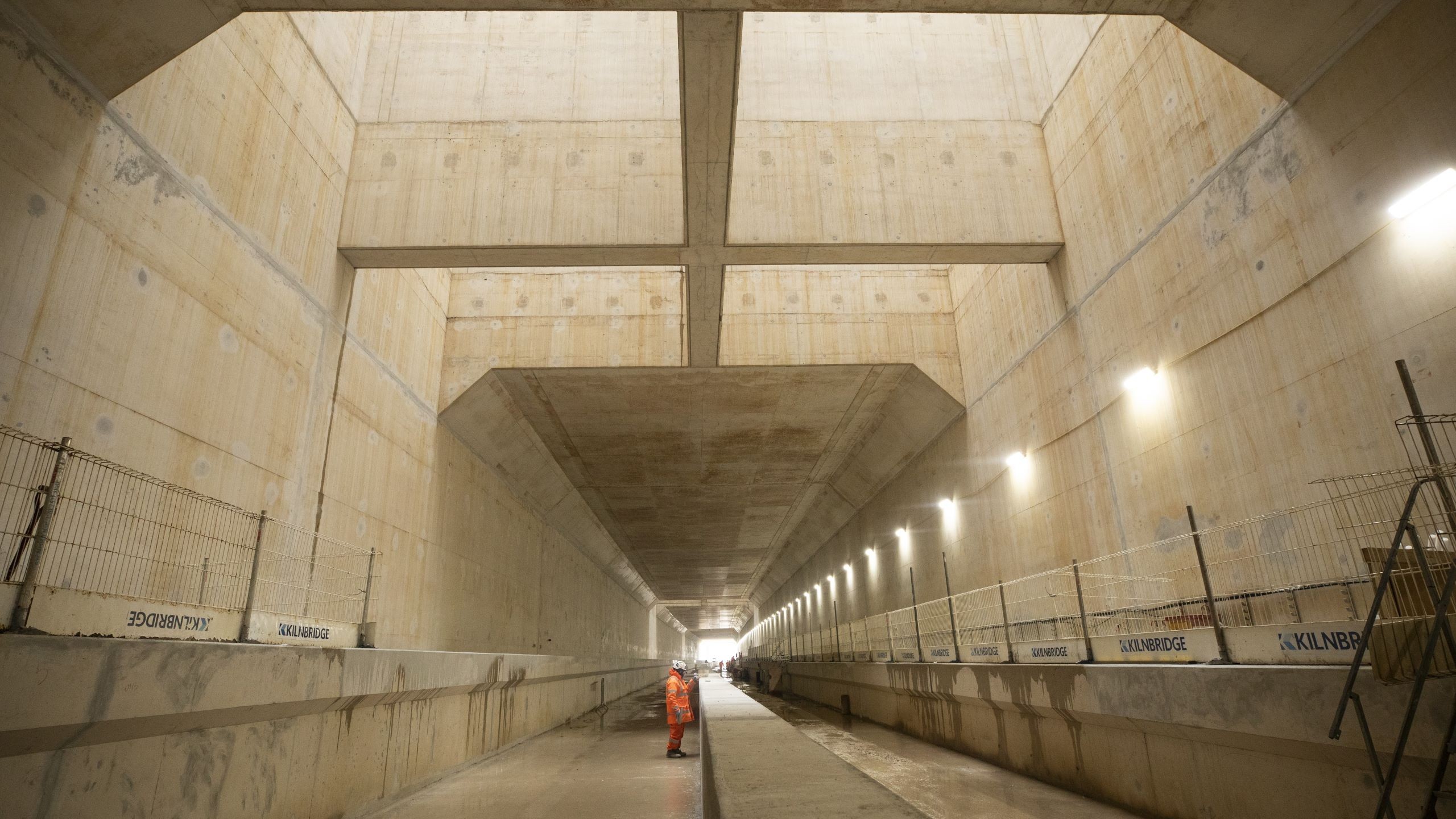 Construction worker inside large concrete tunnel under development Construction worker standing inside a large concrete tunnel under construction.