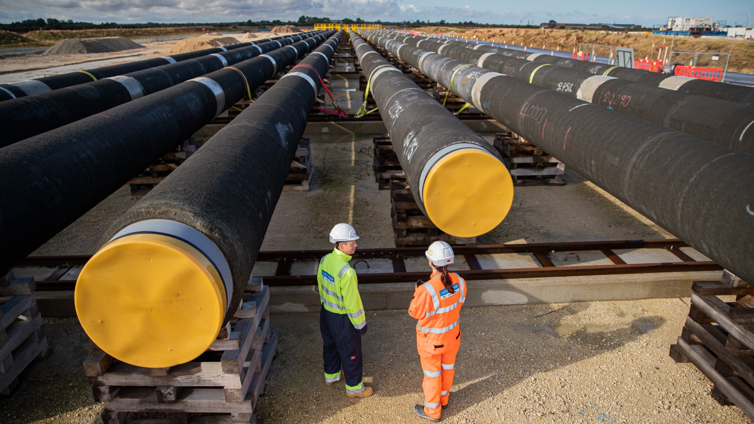 Two workers in safety gear inspect large industrial pipes with yellow caps at a construction site.