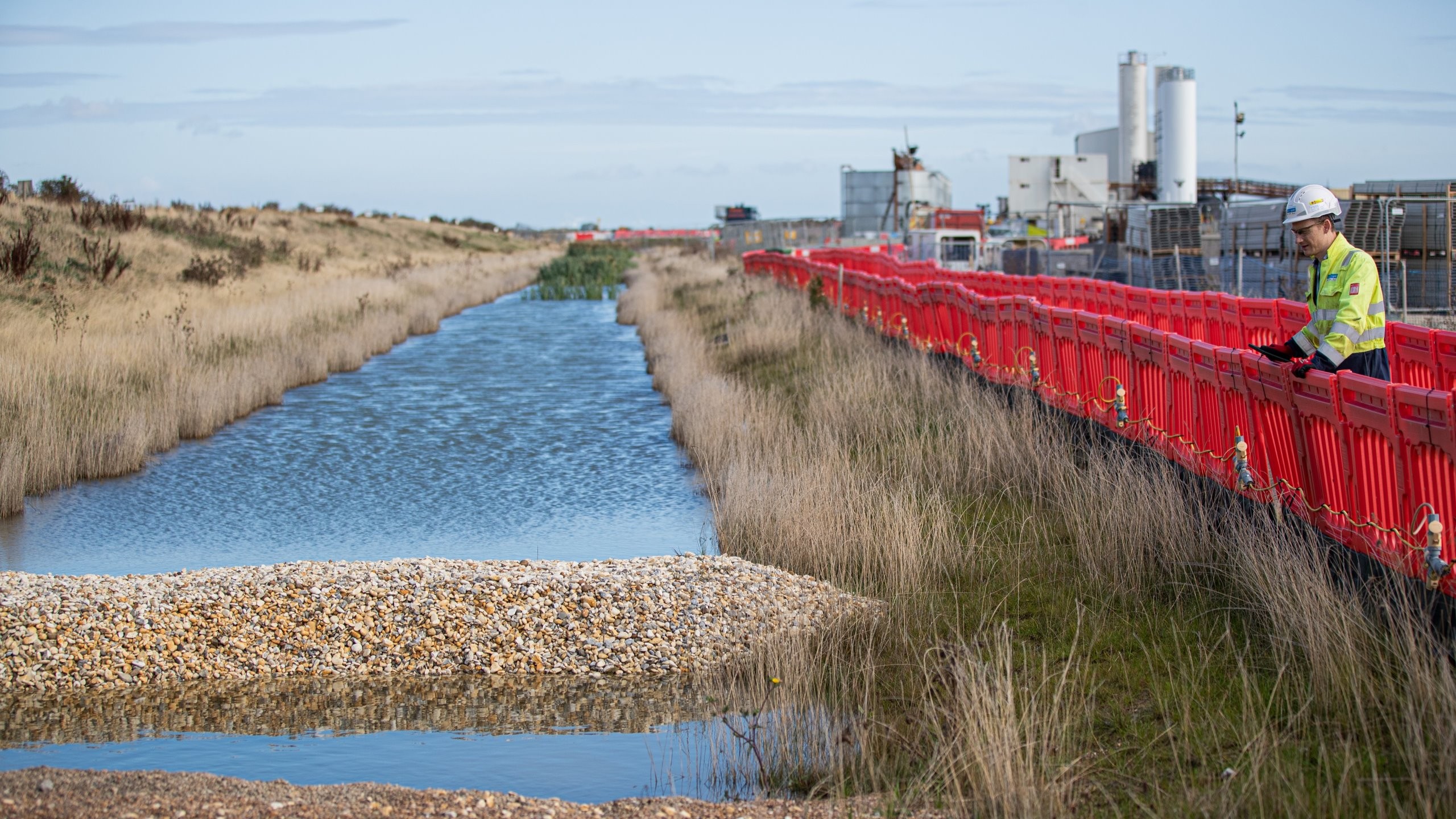 Worker in safety gear inspects waterway with gravel barrier and red fencing nearby.