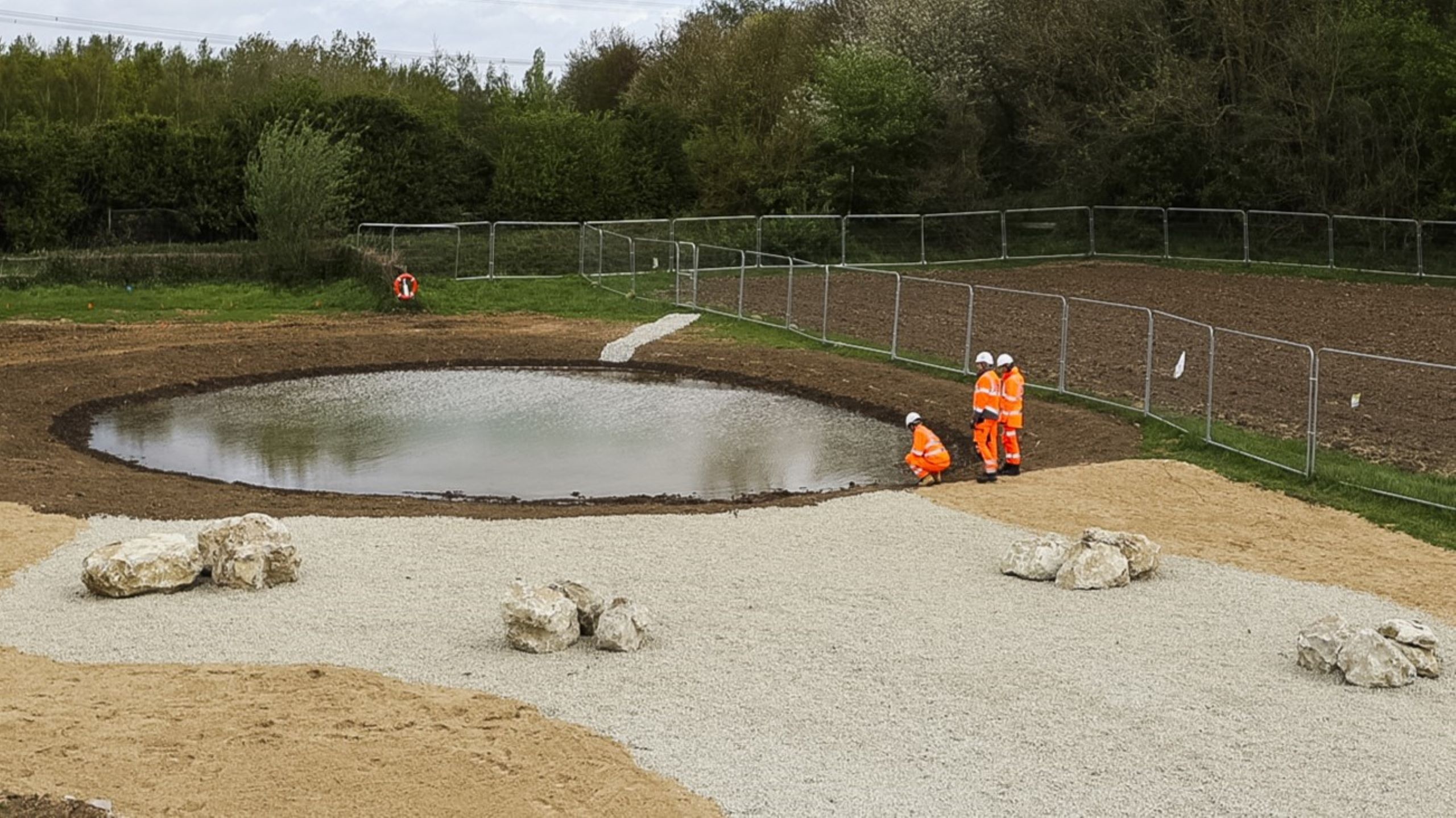 Construction workers in orange safety gear inspect a water retention pond site outdoors.