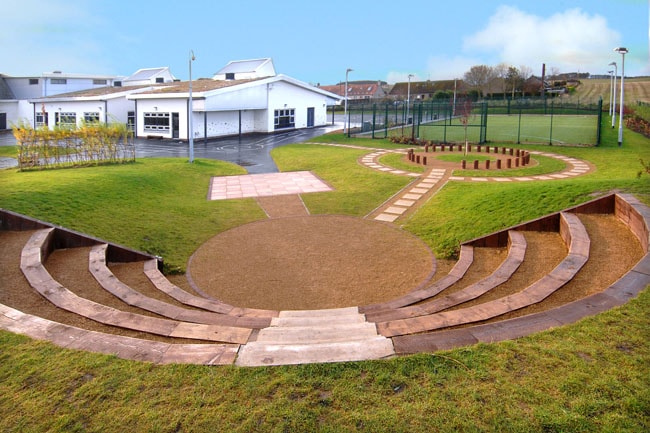 Outdoor amphitheater with seating and sports field in background Outdoor amphitheater with curved wooden benches and a fire pit near a white building.