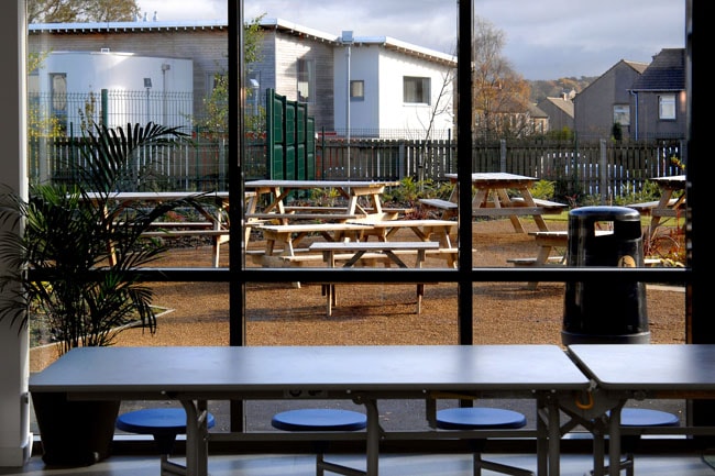 Outdoor picnic tables seen through large window with seating. View of outdoor picnic tables and benches through large glass windows in a school cafeteria.
