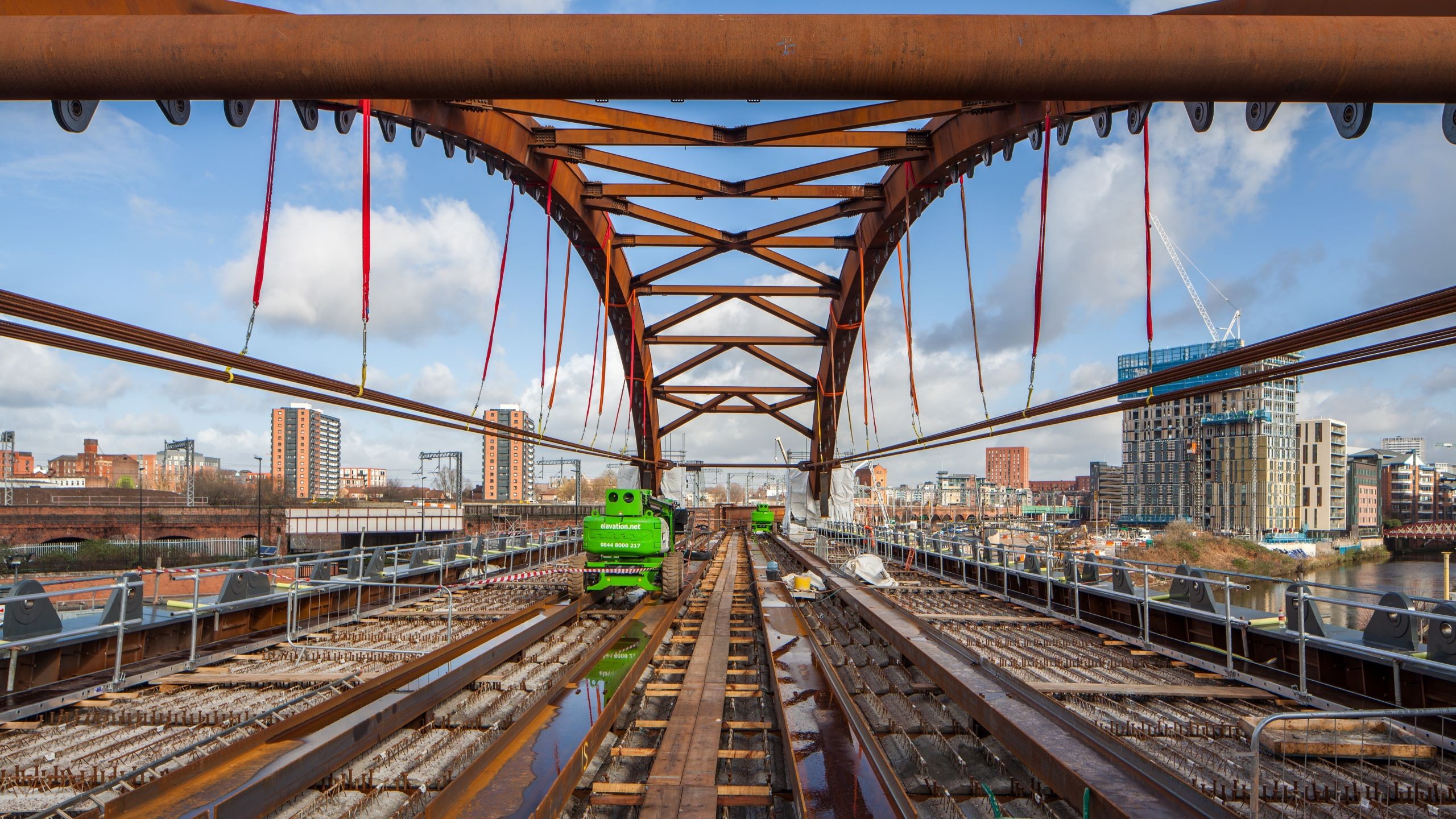 Steel bridge under construction over railway tracks with city buildings in background.