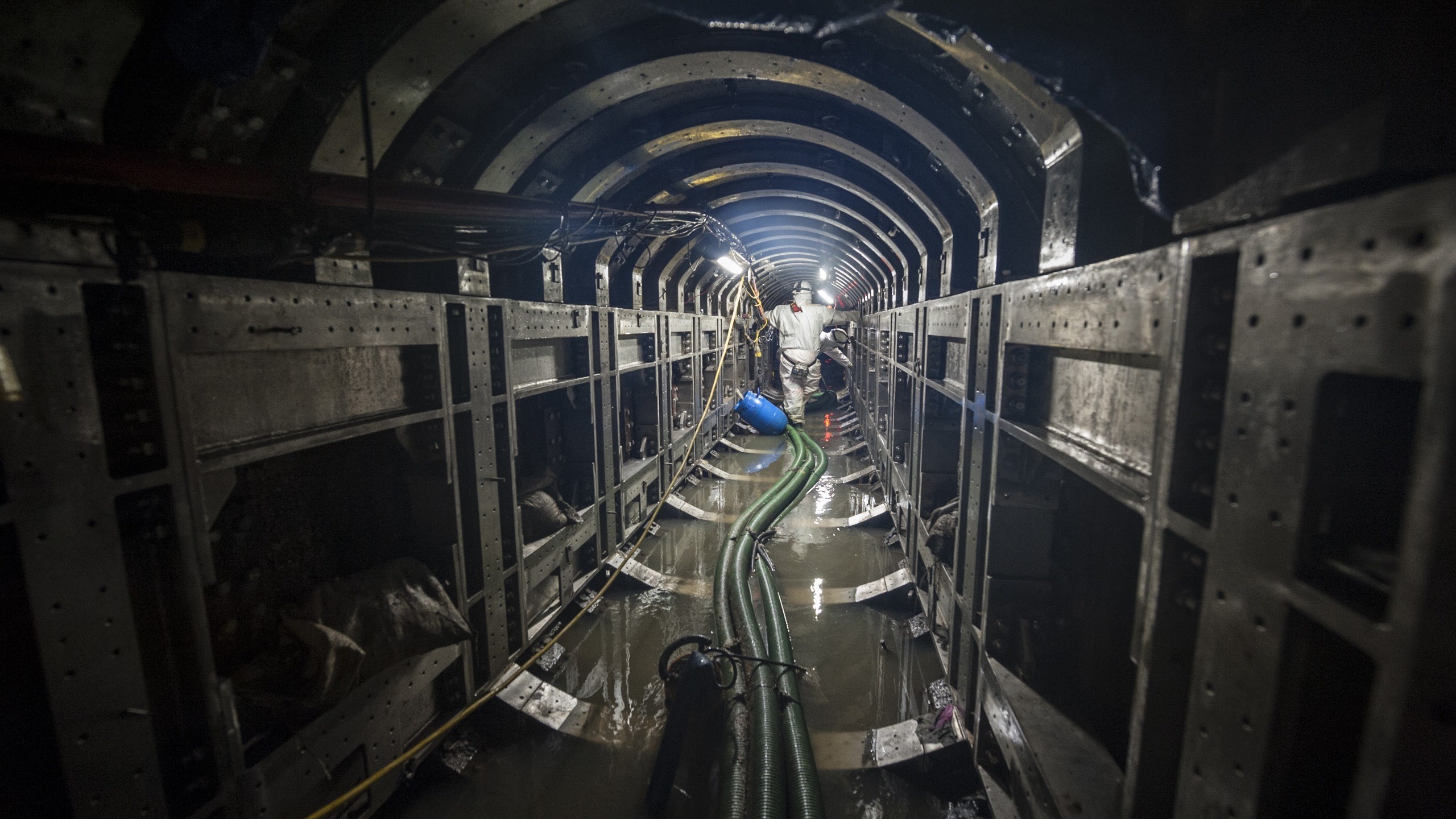 Tunnel Construction with Heavy Machinery and Workers Worker in protective gear walking through a flooded industrial tunnel with pipes.