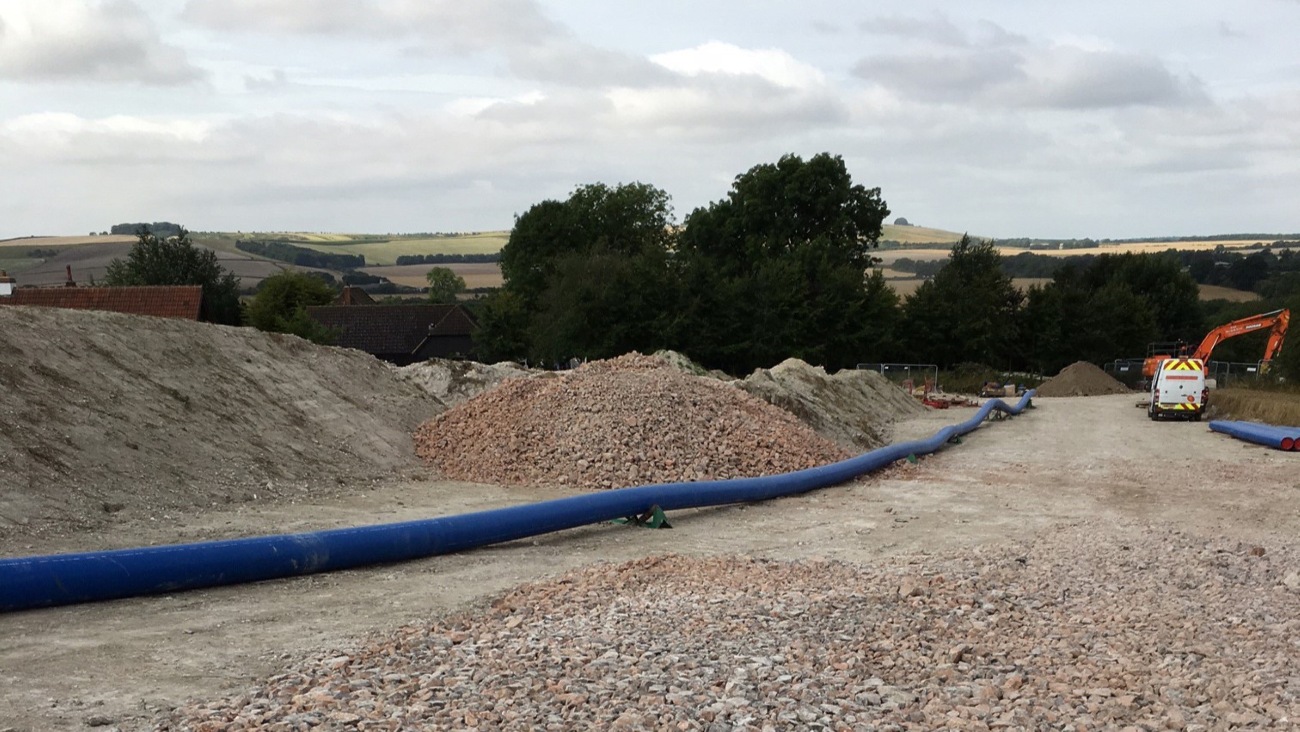 Construction site with blue pipeline, gravel piles, and excavator under cloudy sky.