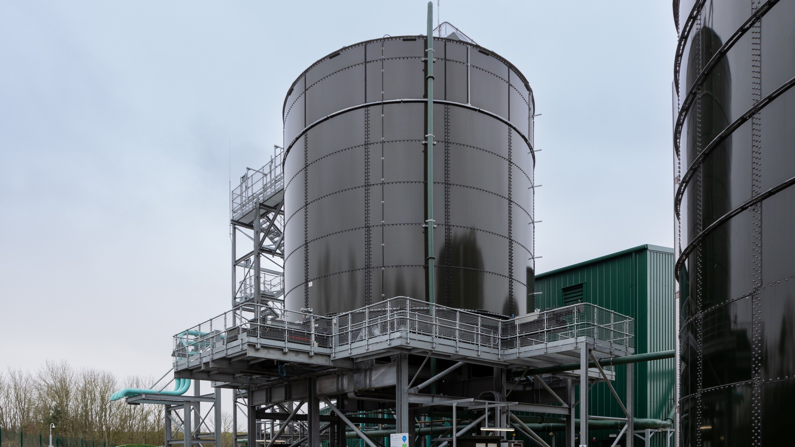 Large industrial storage tank with metal framework and green building under overcast sky.