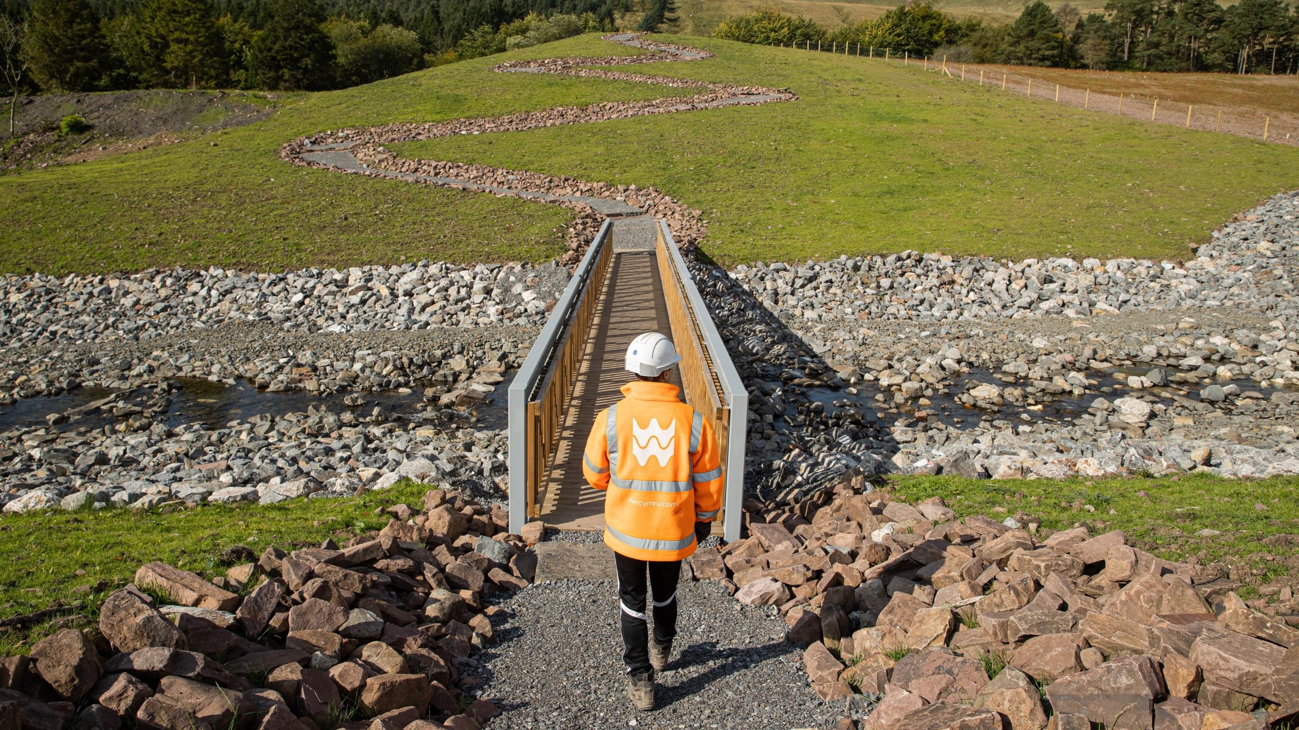 Construction Site Development with Heavy Machinery Worker in orange safety jacket walks on rocky path toward narrow metal bridge over stream.