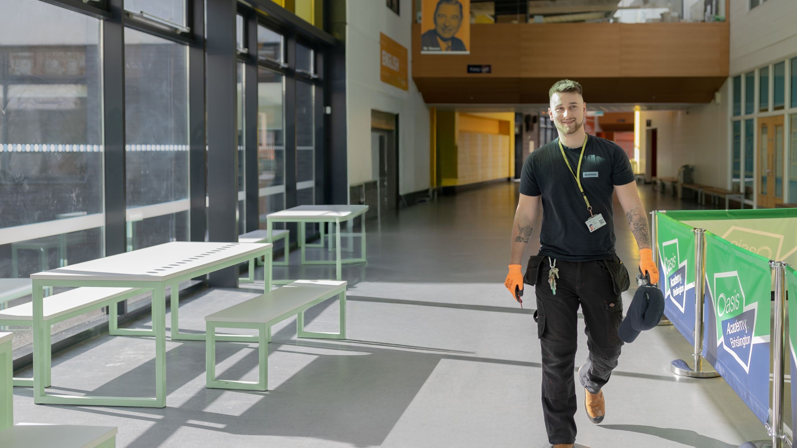 Man in black uniform walking through a bright, modern school hallway with tables.