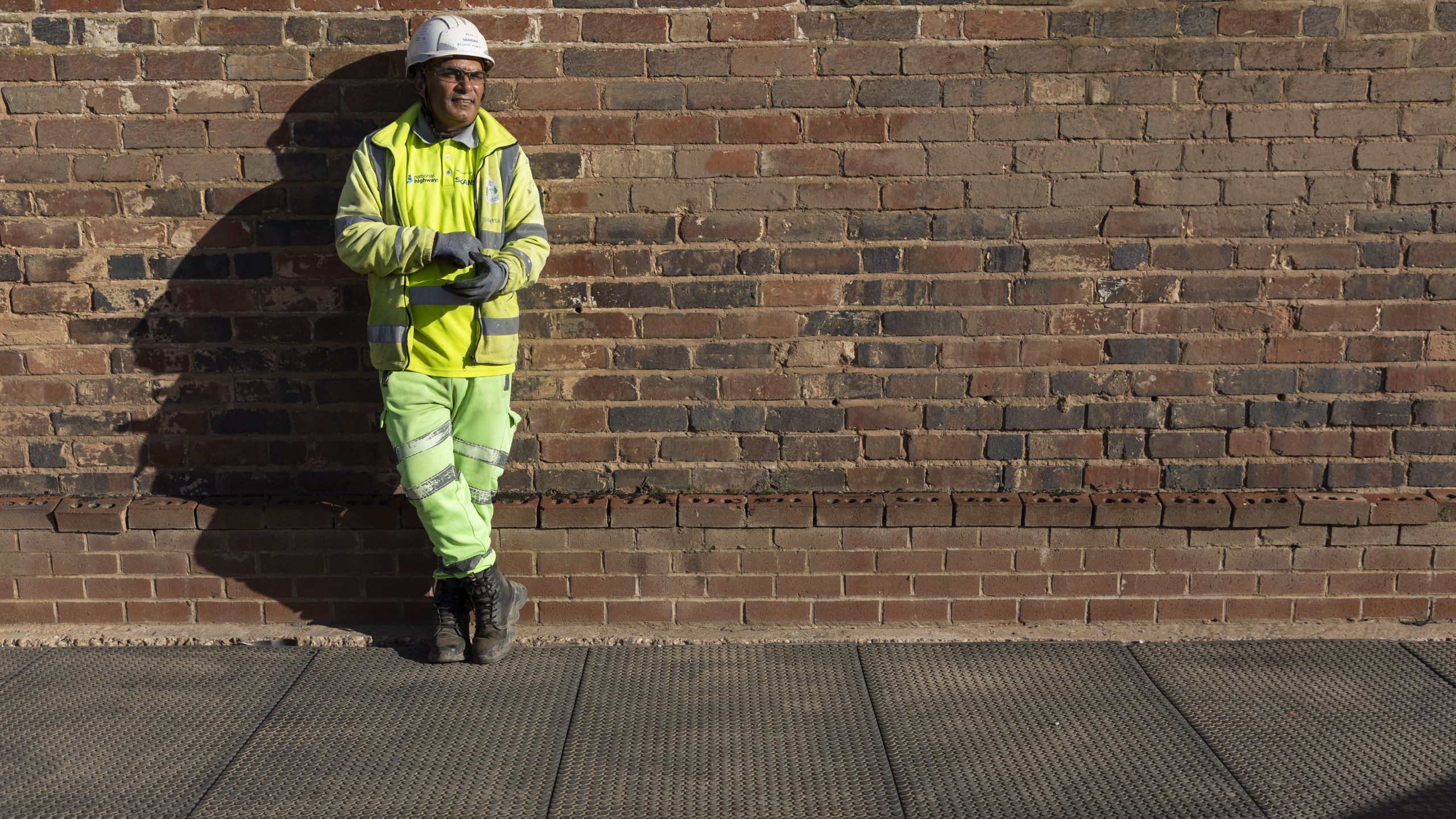 Construction worker in safety gear leaning on brick wall outdoors Construction worker in high-visibility gear and helmet leaning against brick wall outdoors.