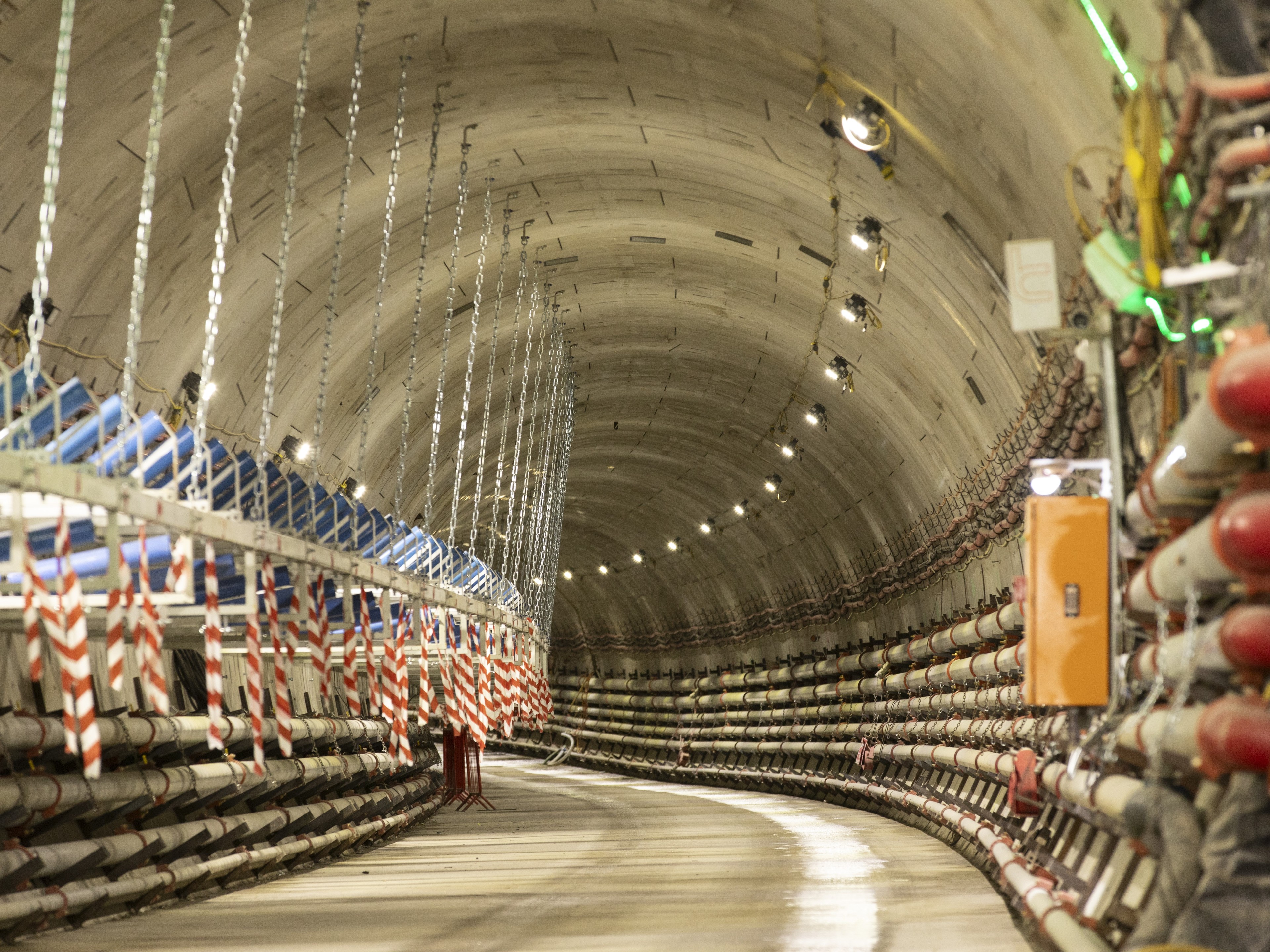 Underground Tunnel with Pipes and Lighting Fixtures Curved underground tunnel with suspended cables and safety barriers, illuminated by overhead lights.