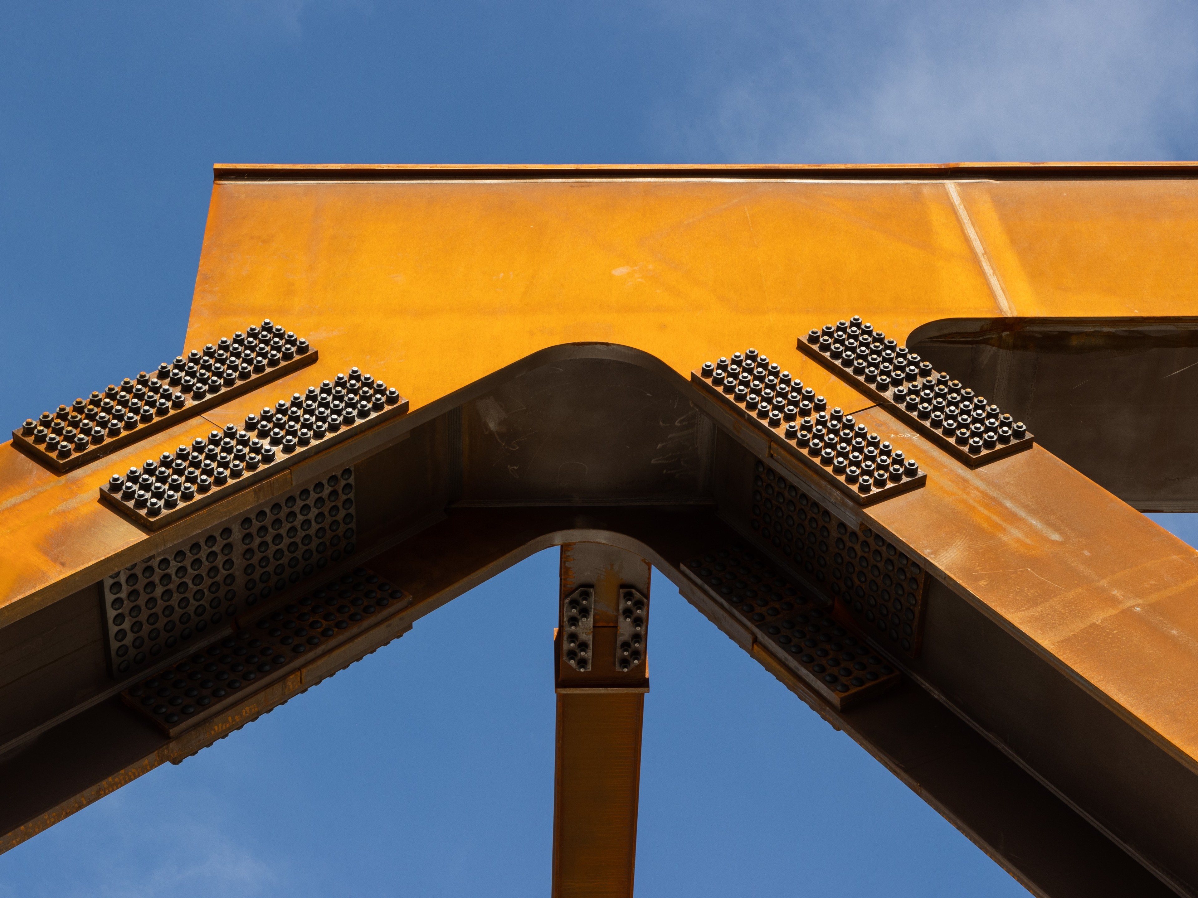Rustic Steel Architecture Against Blue Sky Background Rusty metal beams form a geometric pattern against a clear blue sky, with bolt clusters visible.