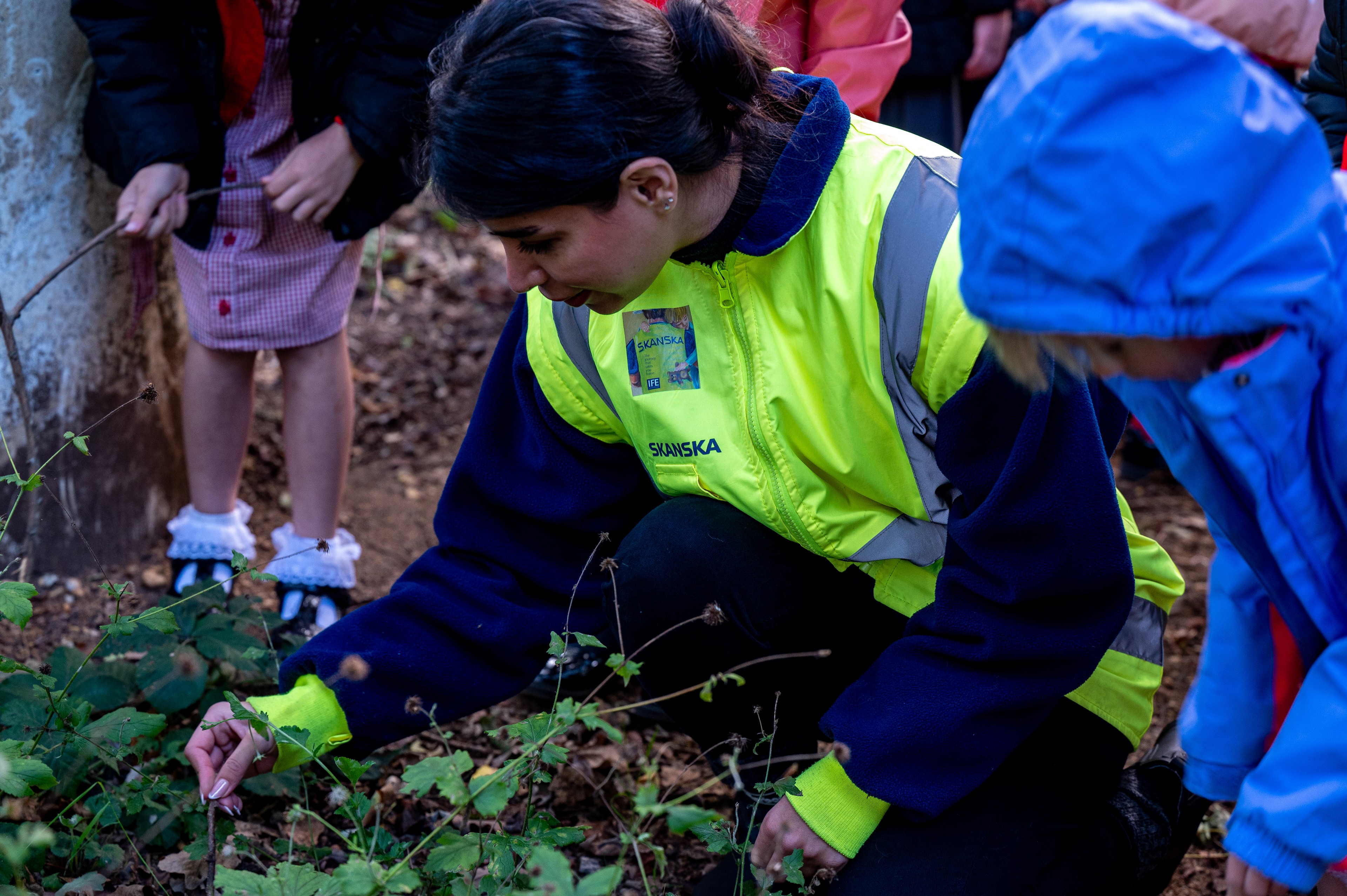 Person wearing a high-visibility jacket gardening outdoors with a child in blue jacket.
