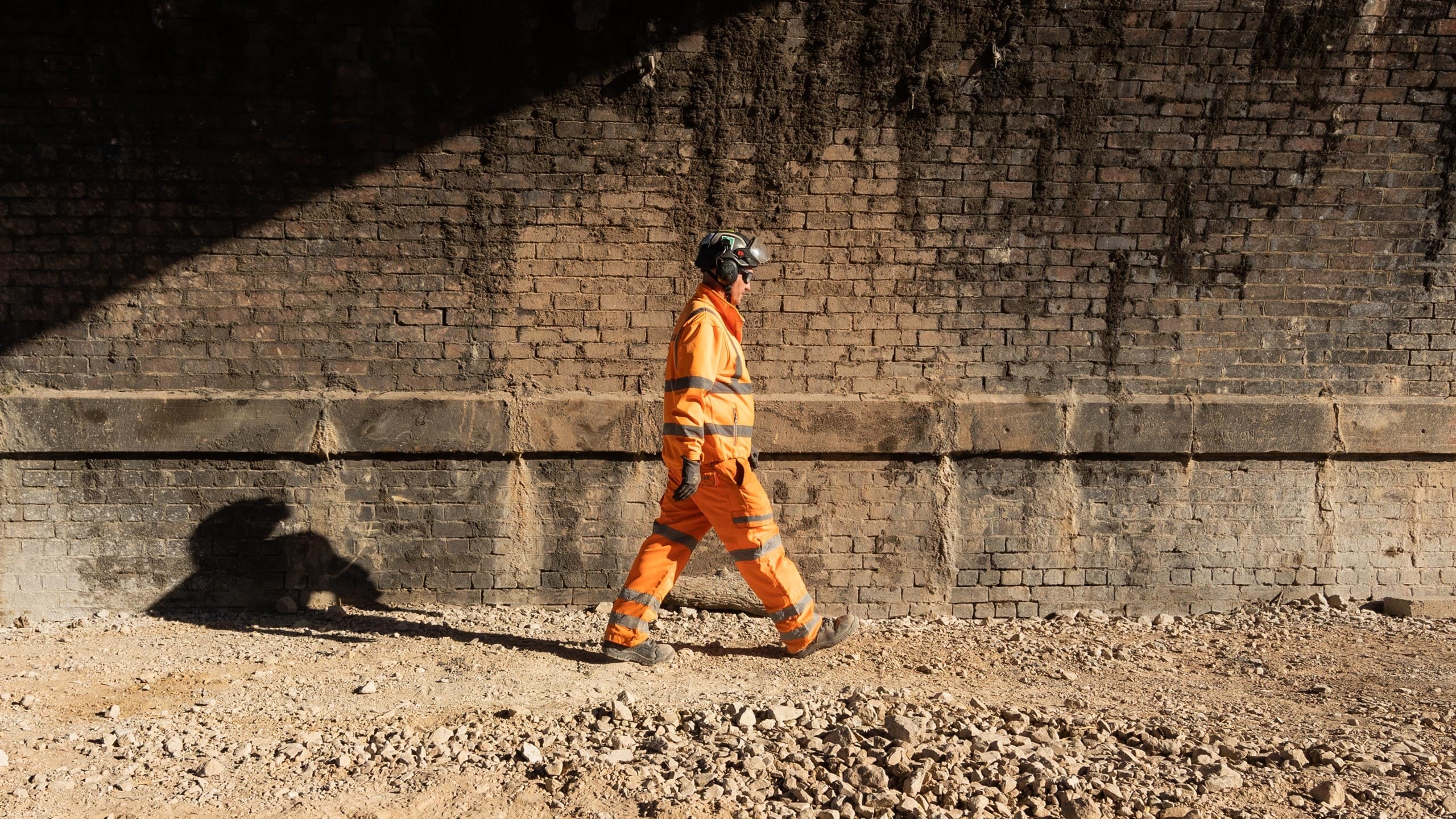 Construction Worker Walking by Brick Wall in Safety Gear A worker in orange safety gear walks along a gravel path beside a brick wall under sunlight.