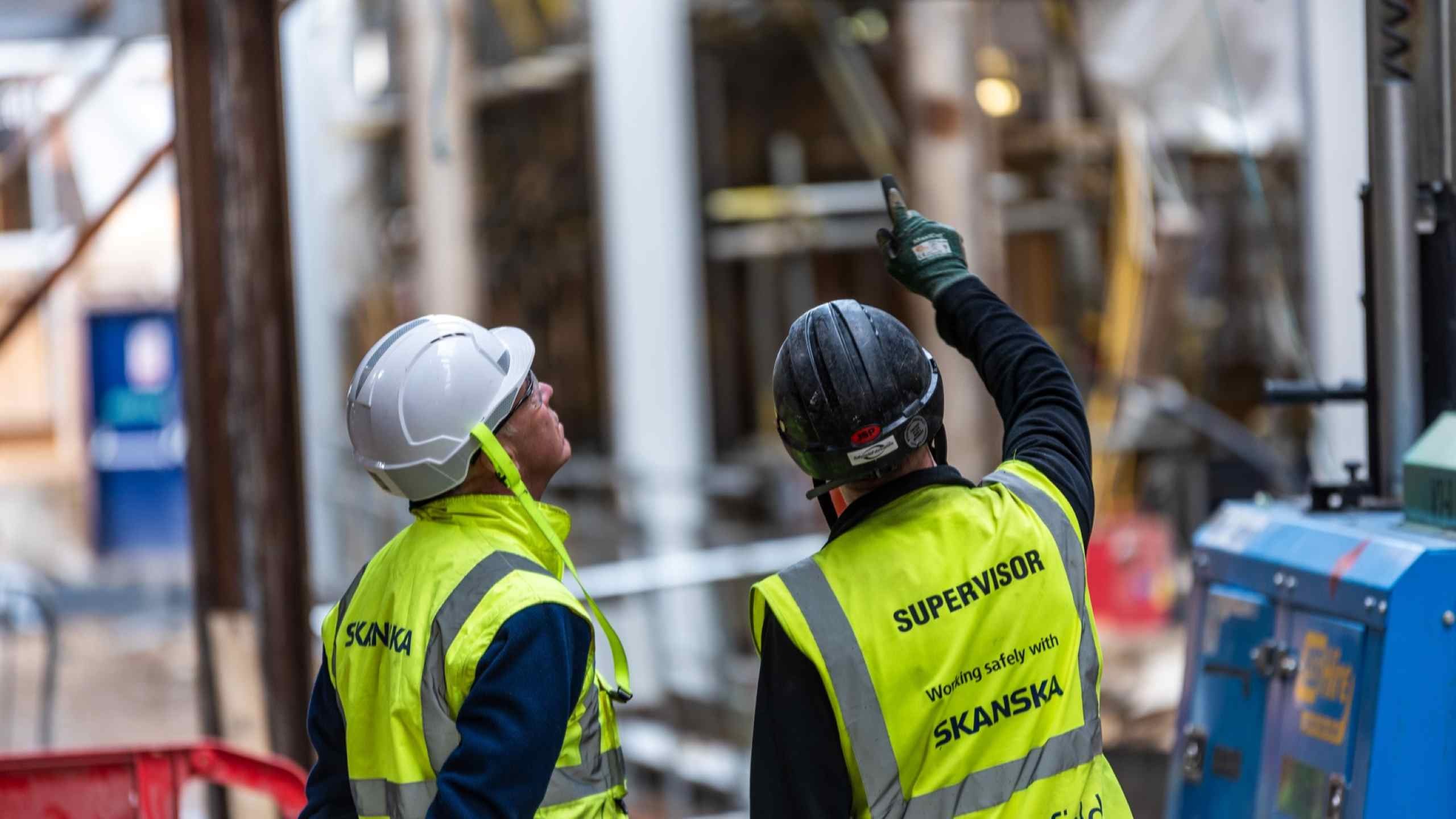 Two construction workers in high-visibility vests and helmets discussing at a busy site.