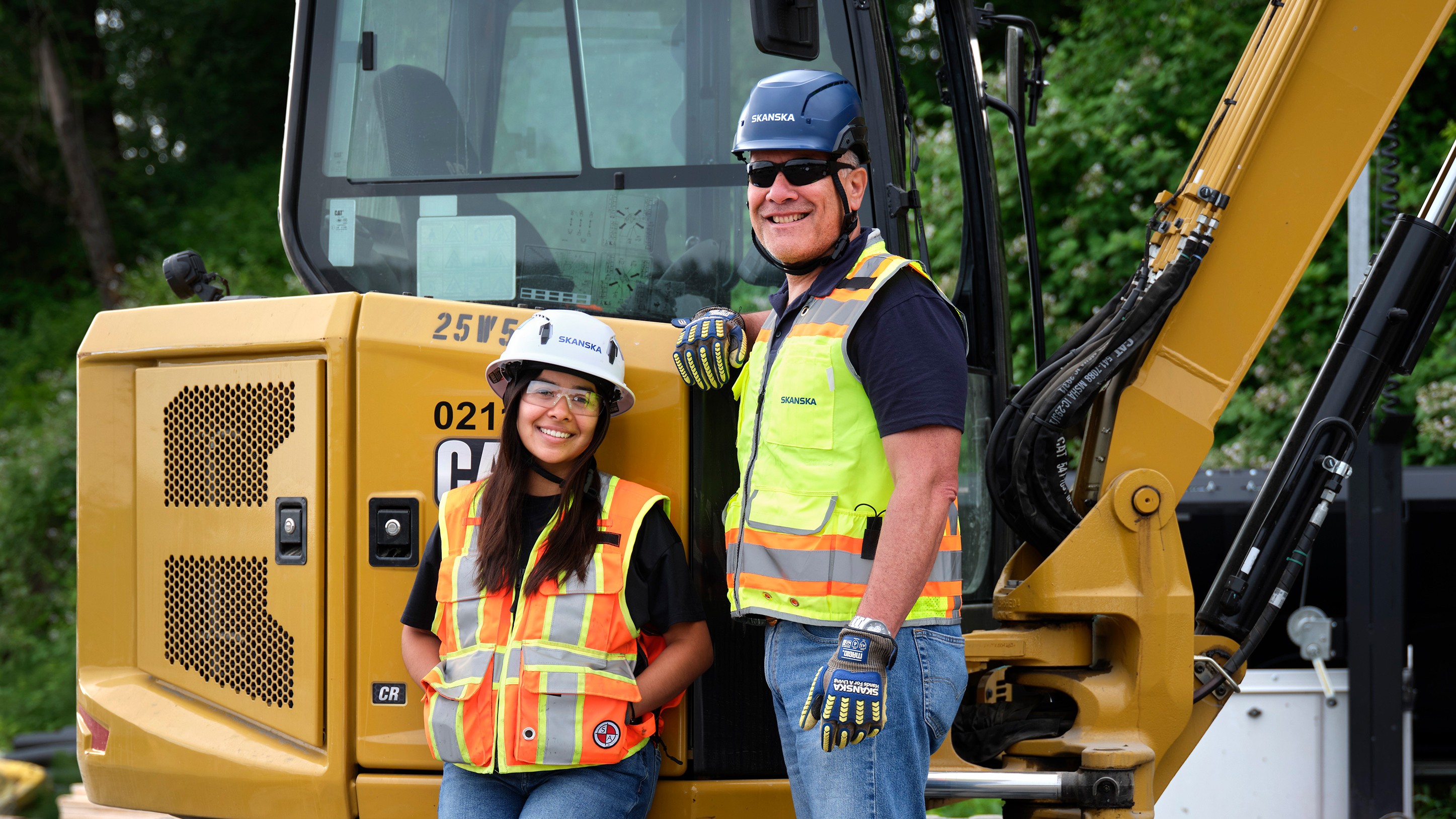 Two construction workers in safety vests and helmets standing by a yellow excavator.