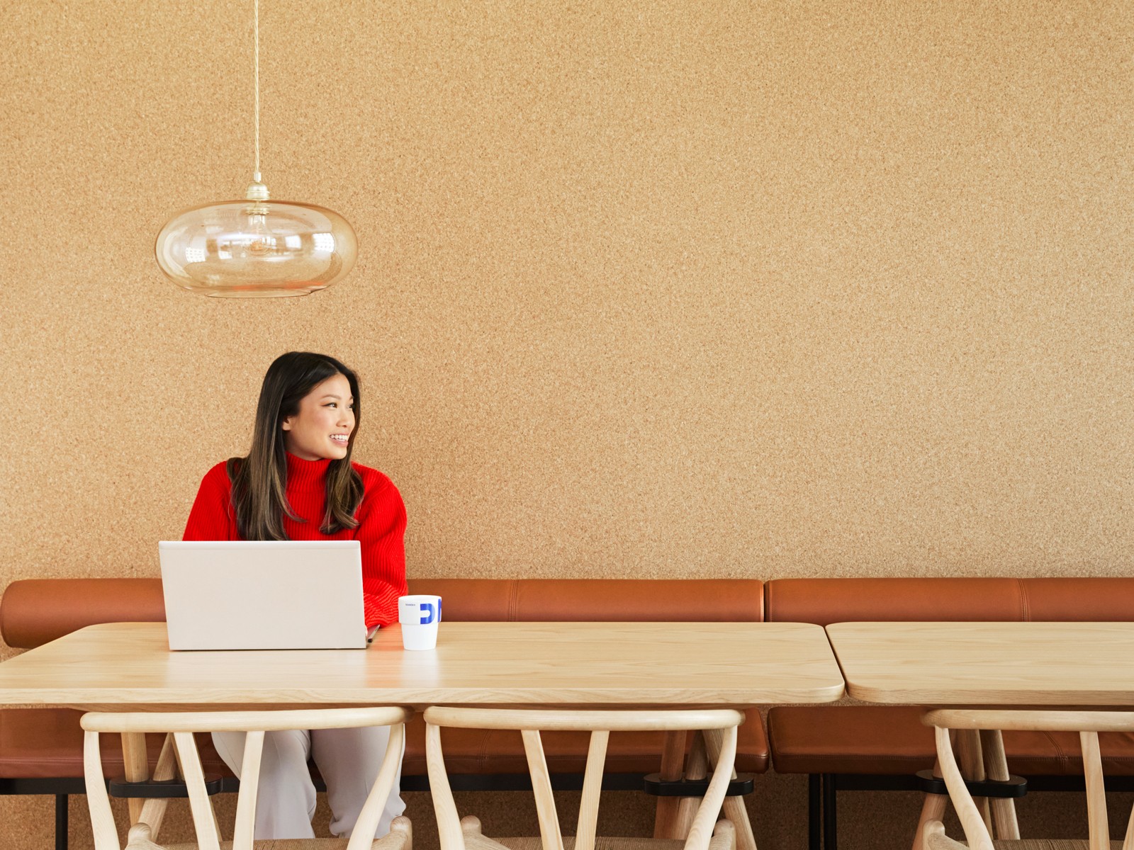 Person Working on a Laptop in a Modern Workspace A person in a red sweater works on a laptop at a light‑wood table in a minimalist workspace, with a pendant light overhead and neutral-toned walls in the background.
