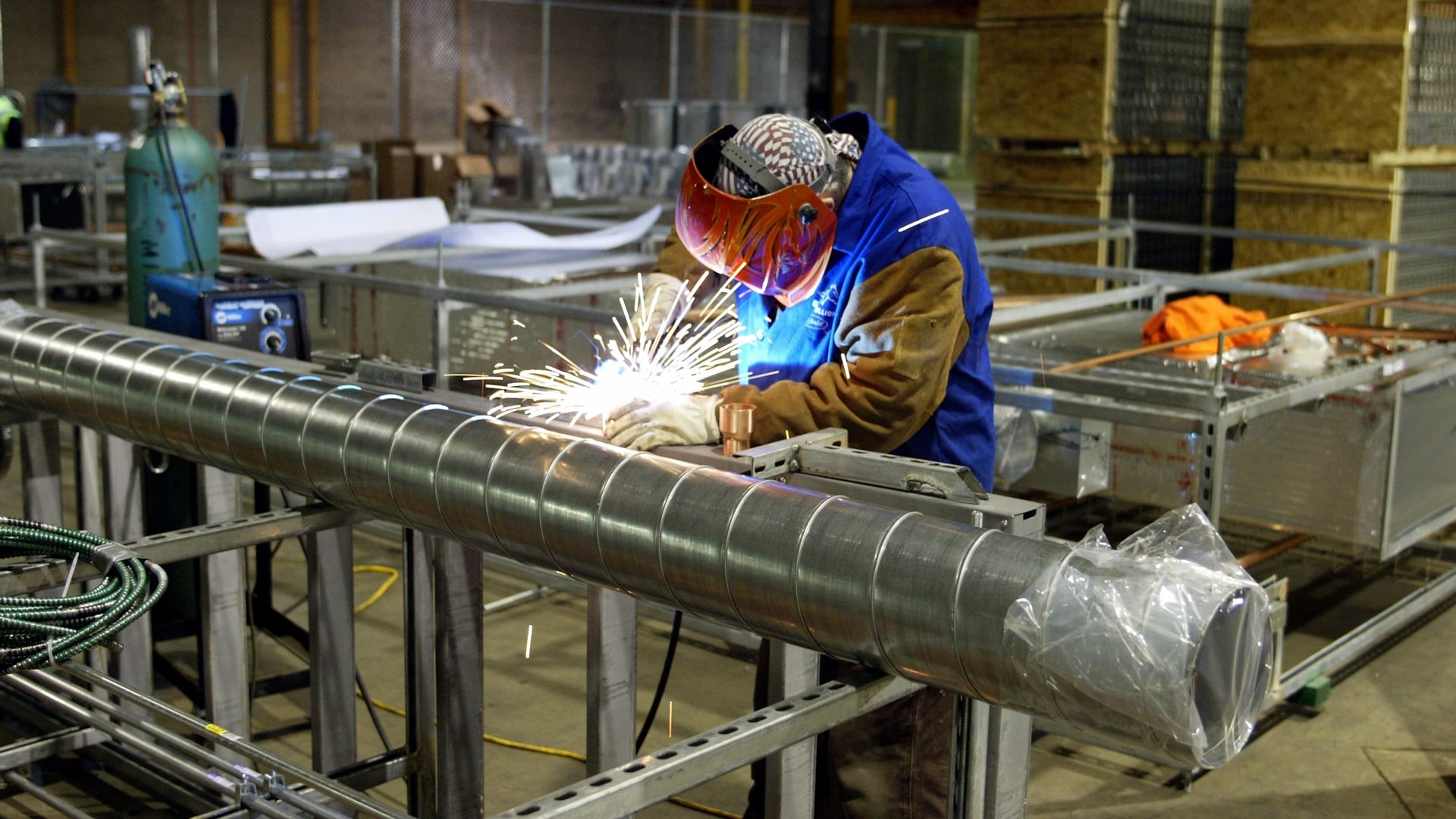 Worker welding metal duct in industrial factory with protective gear and sparks.