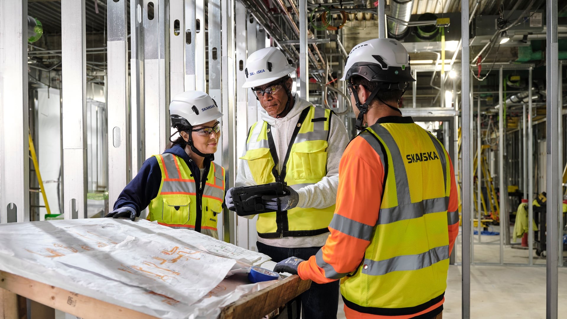 Construction workers in safety gear reviewing blueprints at an indoor building site.