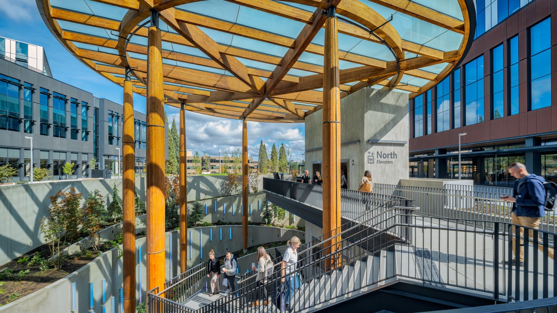 Modern building entrance with wooden columns and glass canopy, featuring people walking on stairs.
