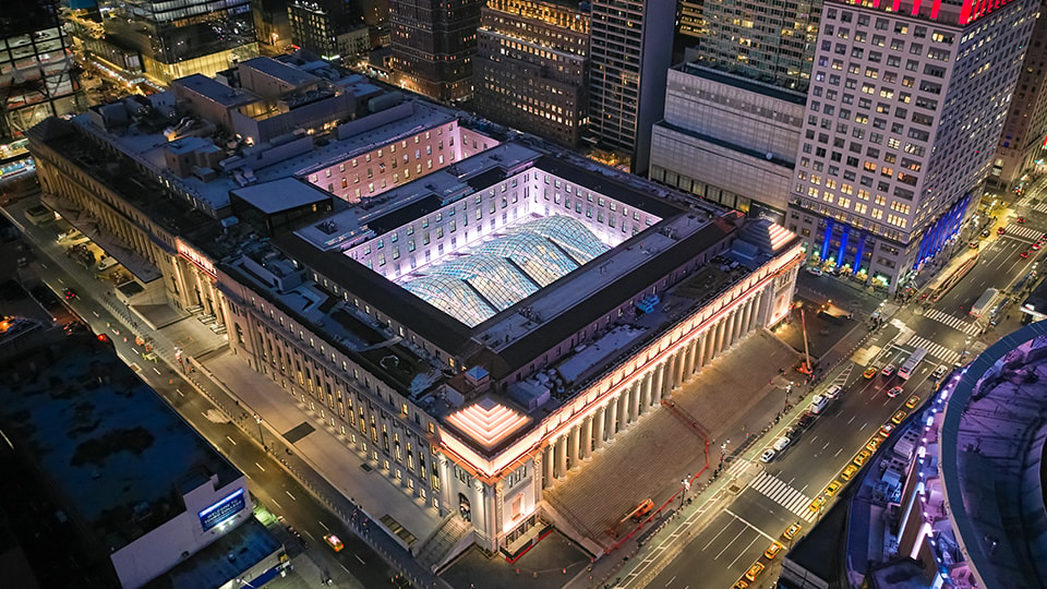 Aerial night view of a large illuminated urban building with surrounding city streets.