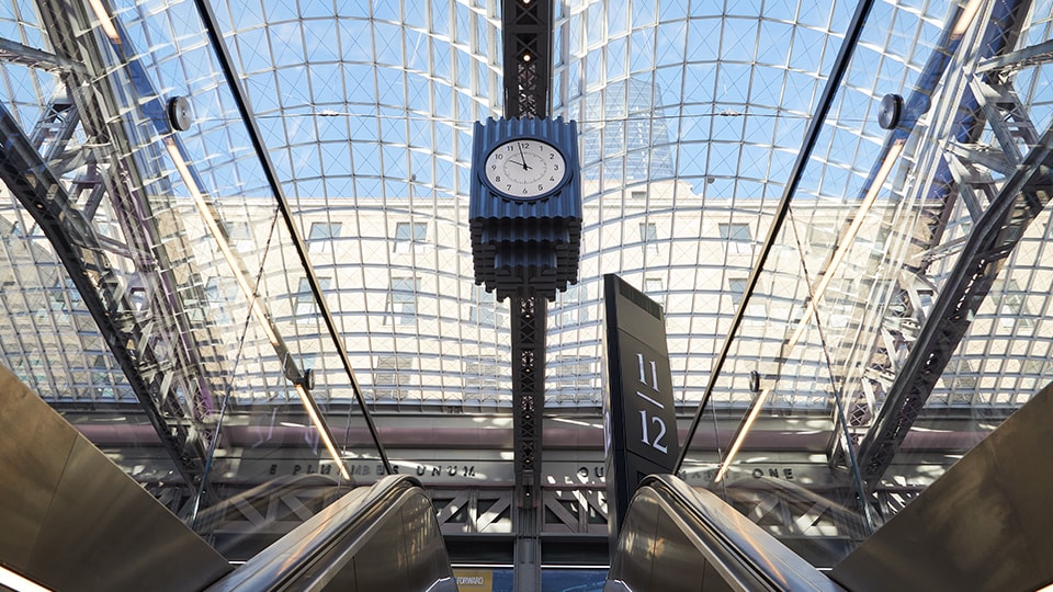 Modern train station interior with glass ceiling, escalators, and a central clock.