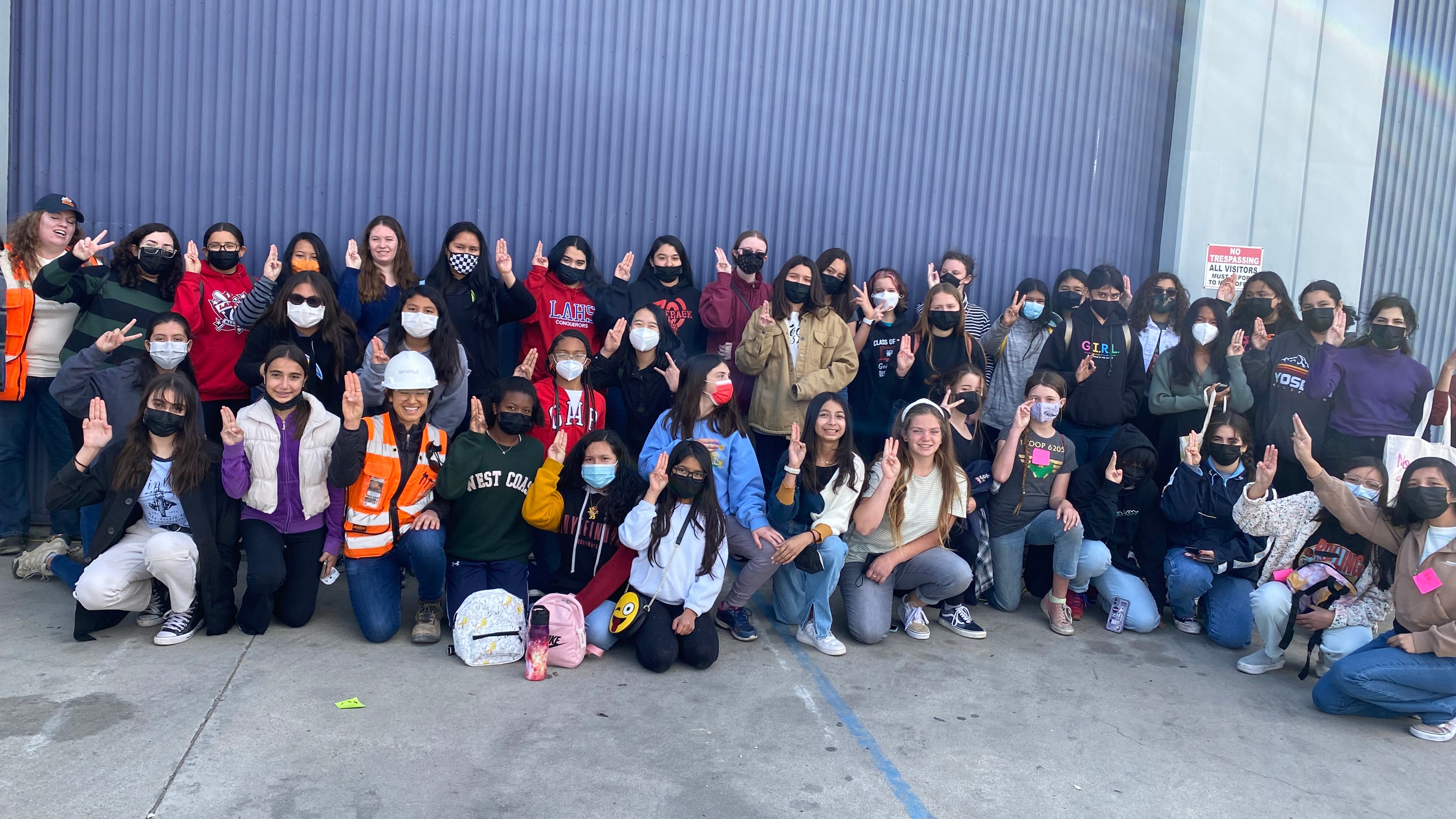 Diverse group of young adults posing outdoors in front of a corrugated metal wall.