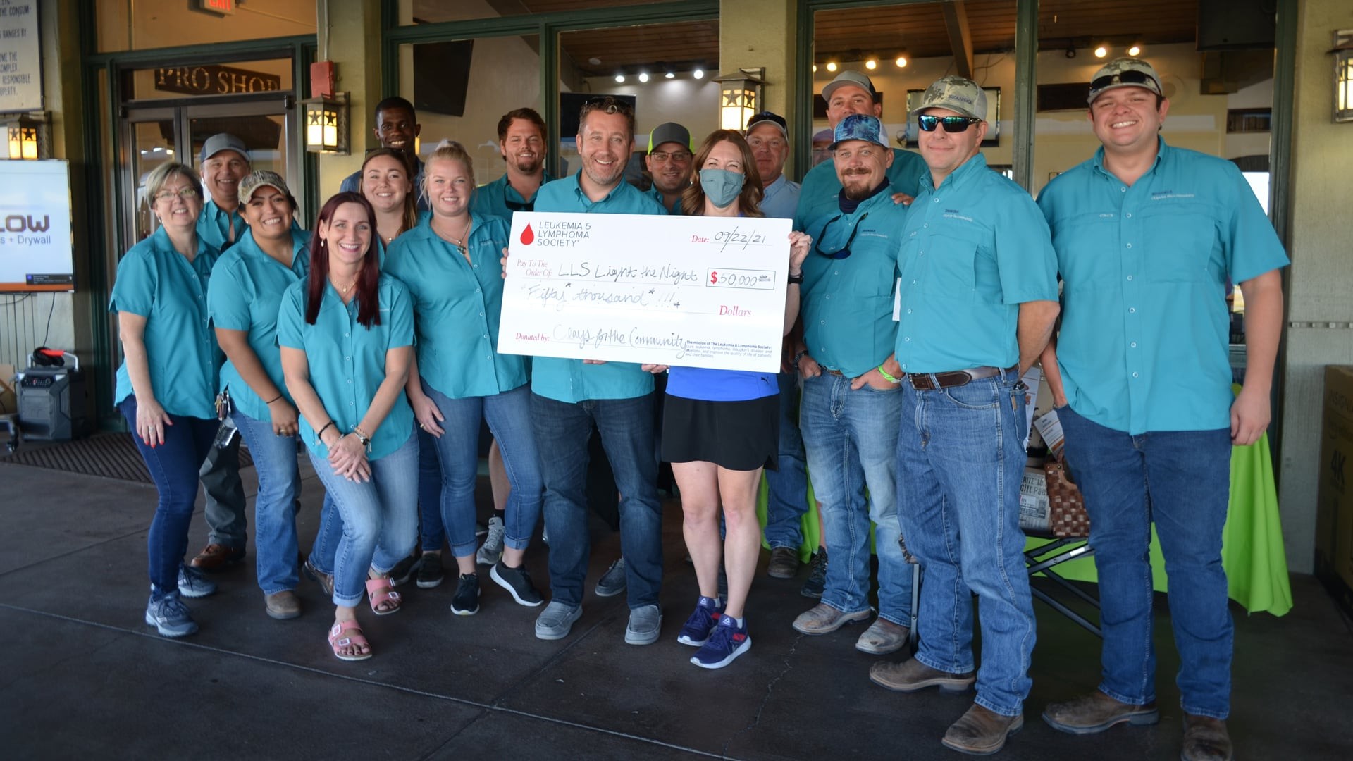 Group of people in teal shirts holding a large donation check indoors at a community event.