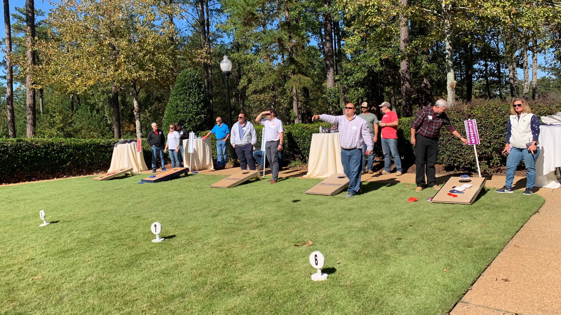 People playing cornhole outdoors on a sunny day with trees and green grass background.