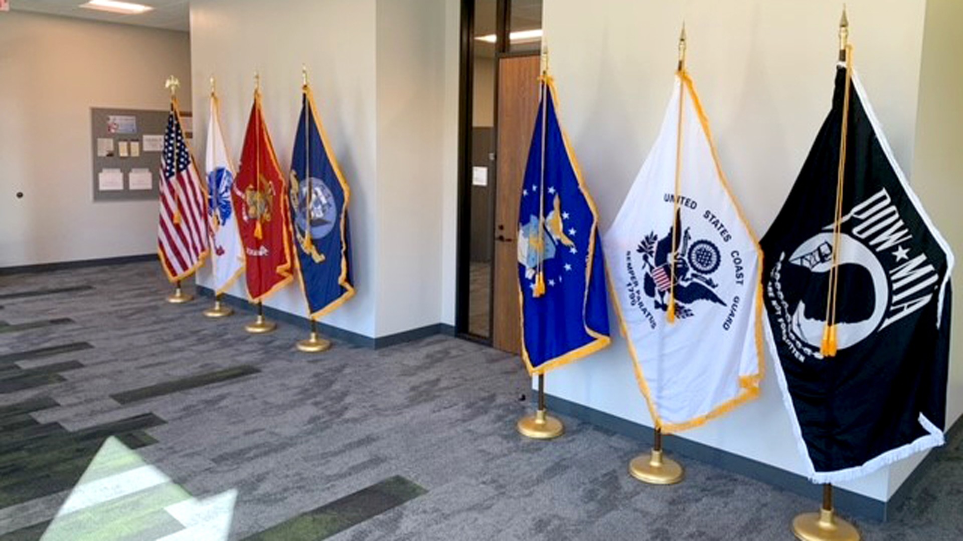 Row of U.S. military and POW/MIA flags displayed on stands in a bright indoor space.