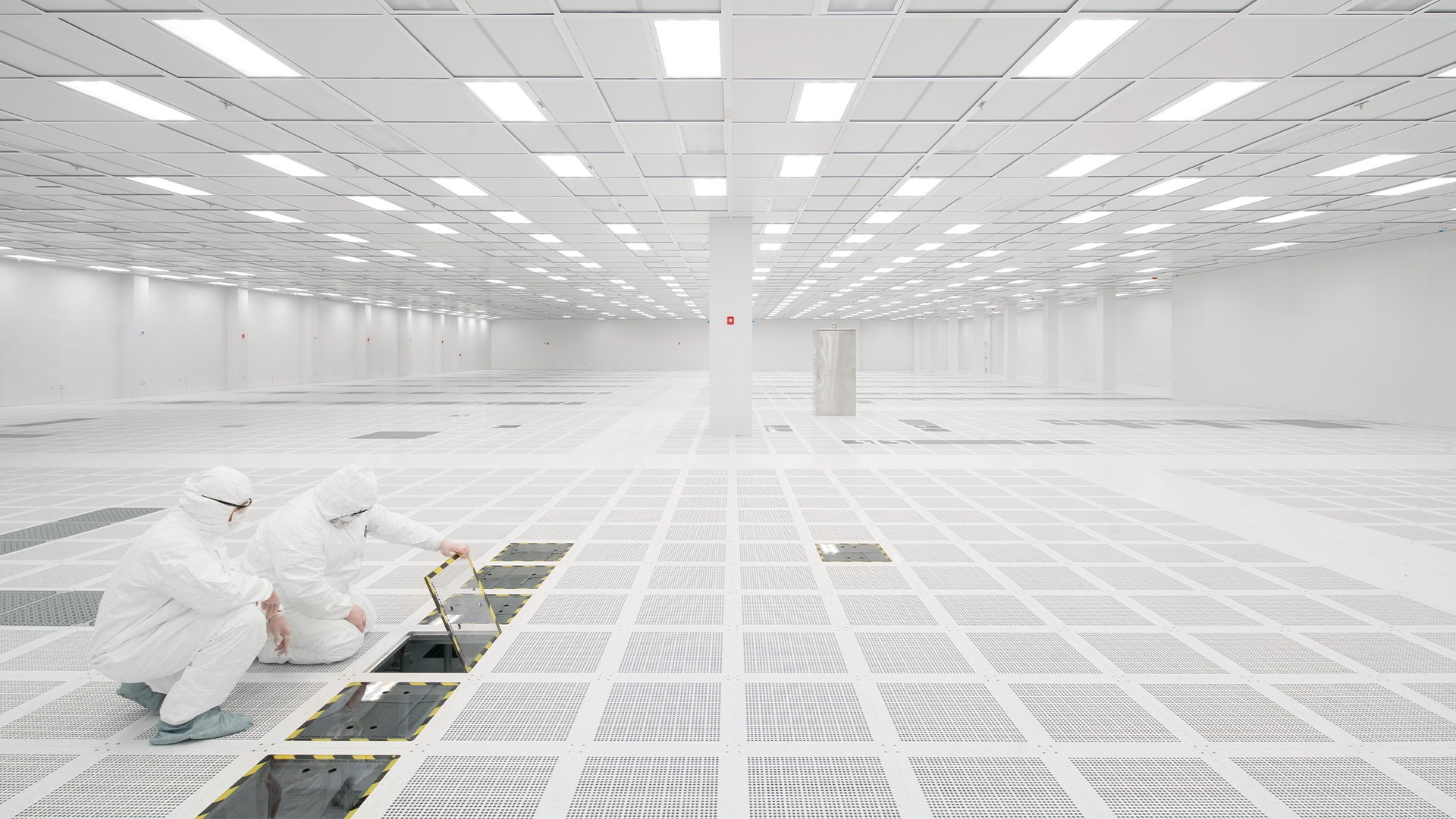 Two workers in protective suits examine an open panel in a vast, pristine cleanroom environment.