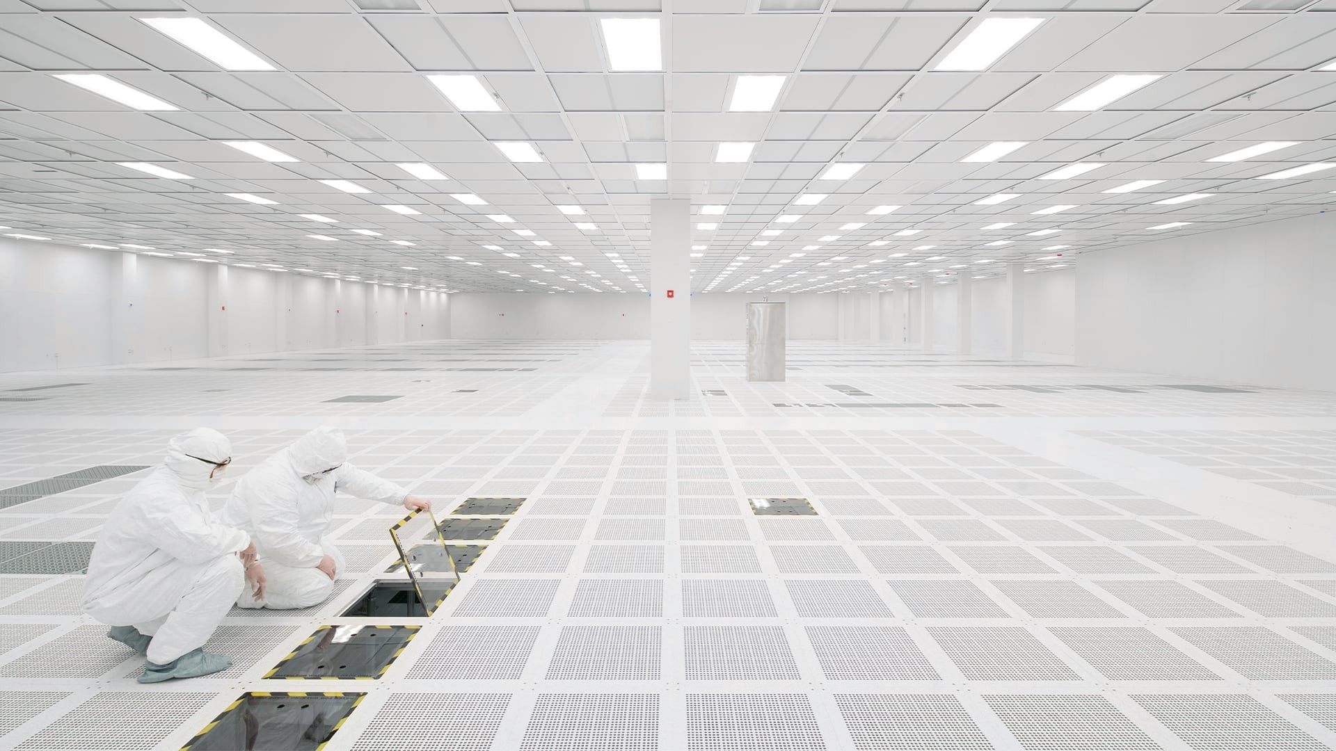Two workers in protective suits examine an open panel in a vast, pristine cleanroom environment.