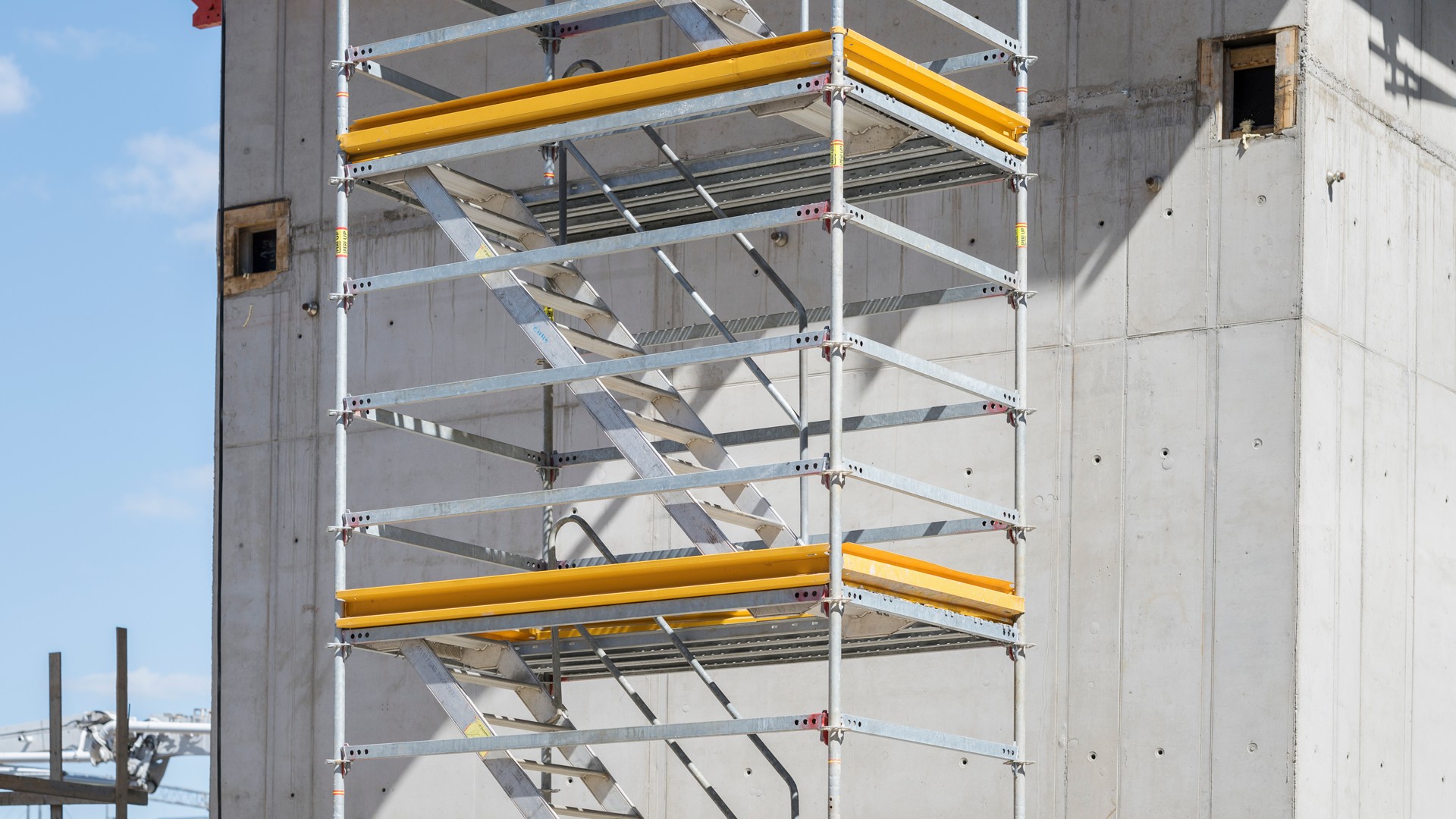 Metallinen teline keltaisilla turvakaiteilla keskeneräisen betonirakennuksen vieressä.