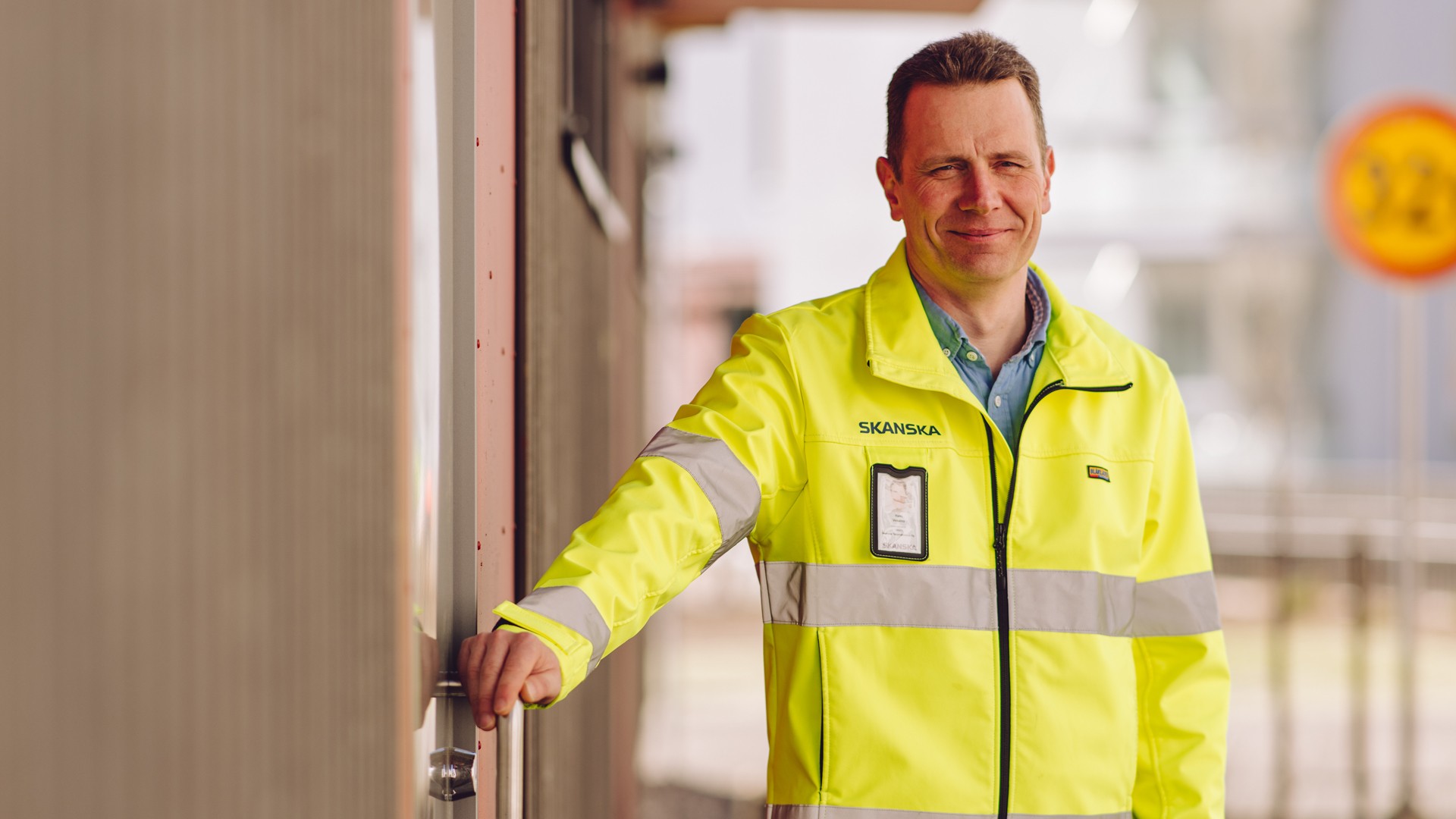 A person in a high-visibility Skanska jacket stands at a door, emphasizing safety and professionalism.