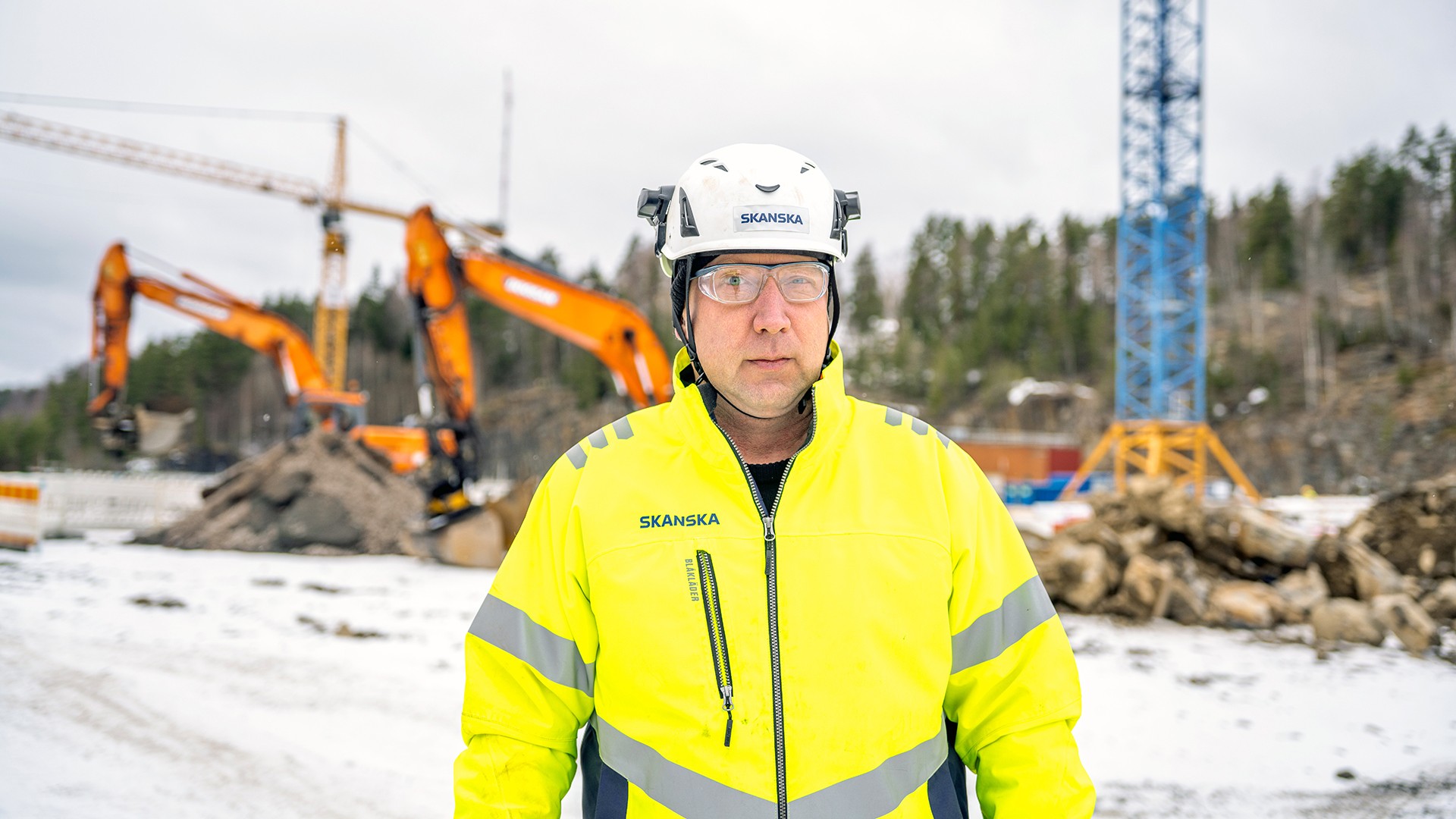 A construction worker in a high-visibility jacket stands at a snowy construction site with machinery.