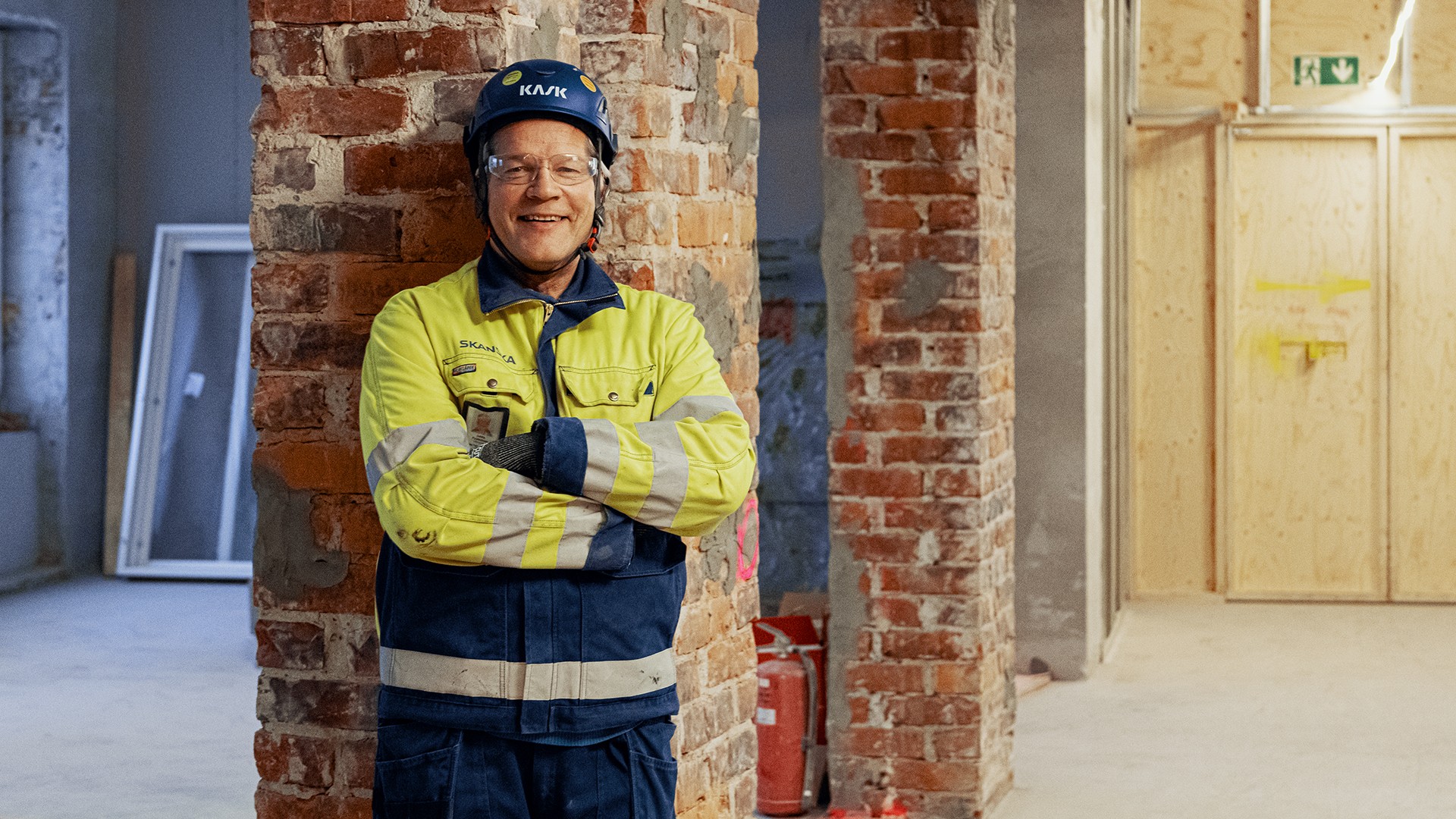 Rakennustyöntekijä suojavarusteissa seisoo käsivarret ristissä tiiliseinän vieressä sisätiloissa.
