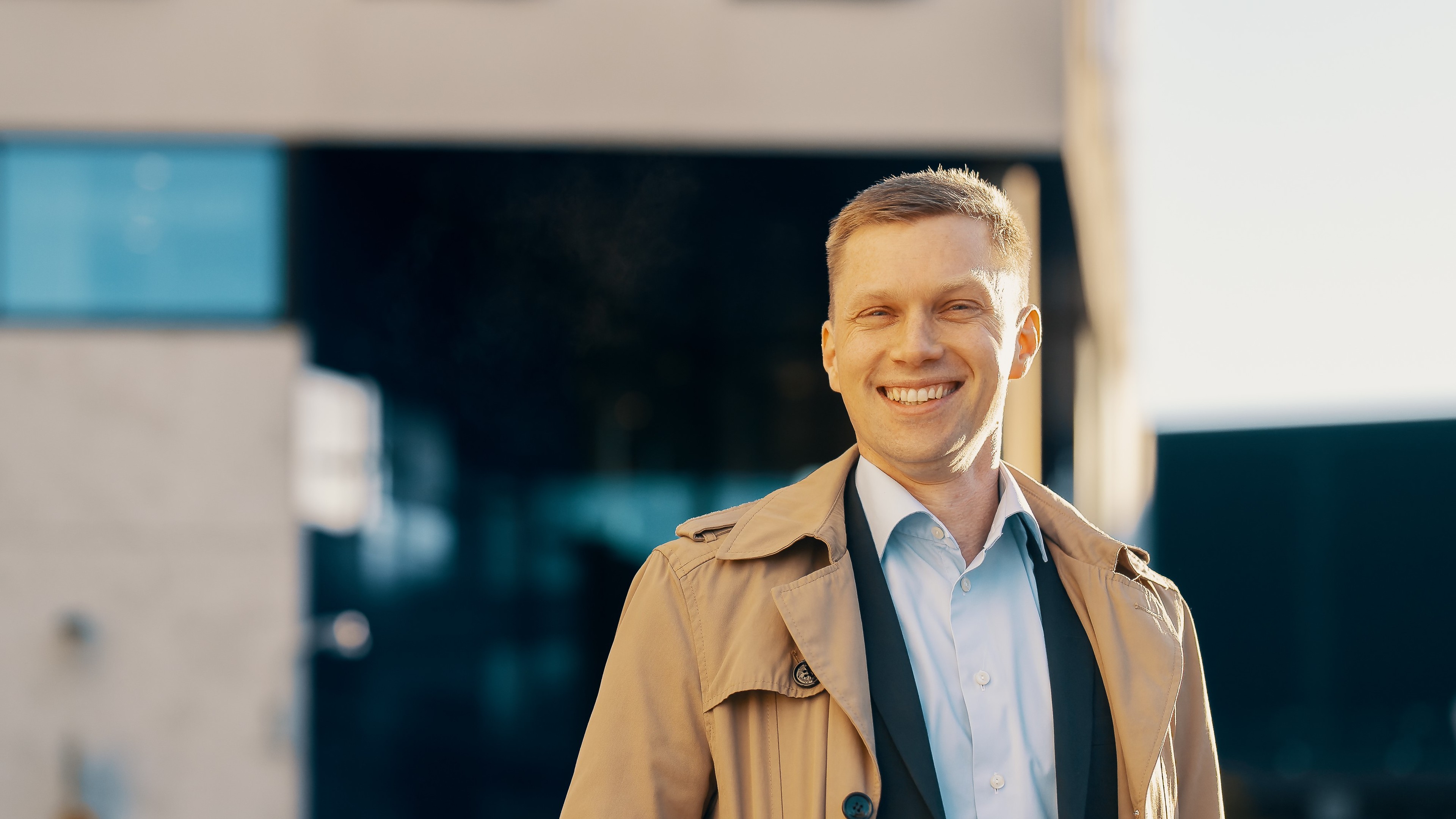 Man in a beige trench coat and blue shirt standing outside a modern building on a sunny day.