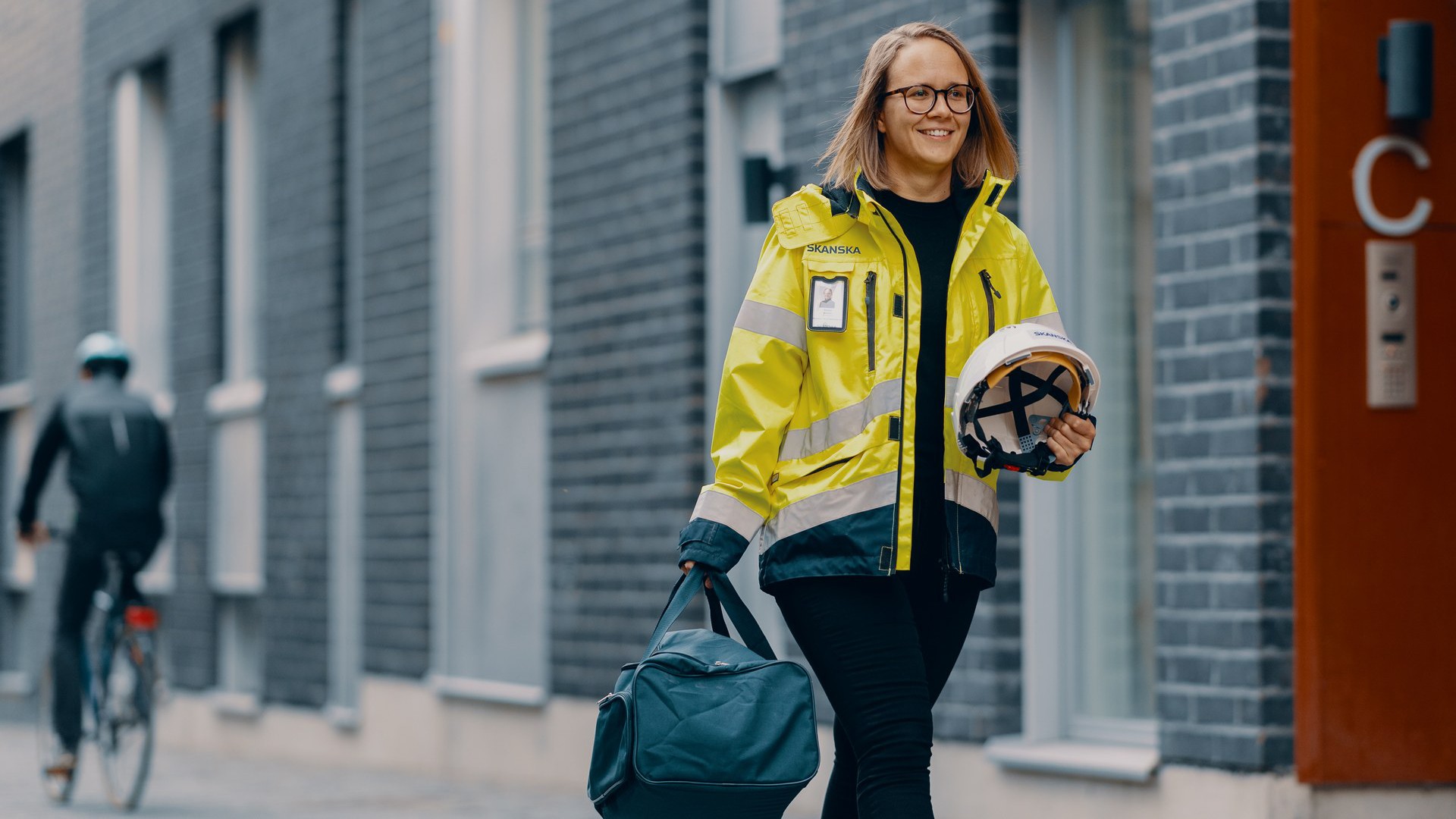 A person in a reflective yellow jacket walks outside a building carrying a helmet and a bag.
