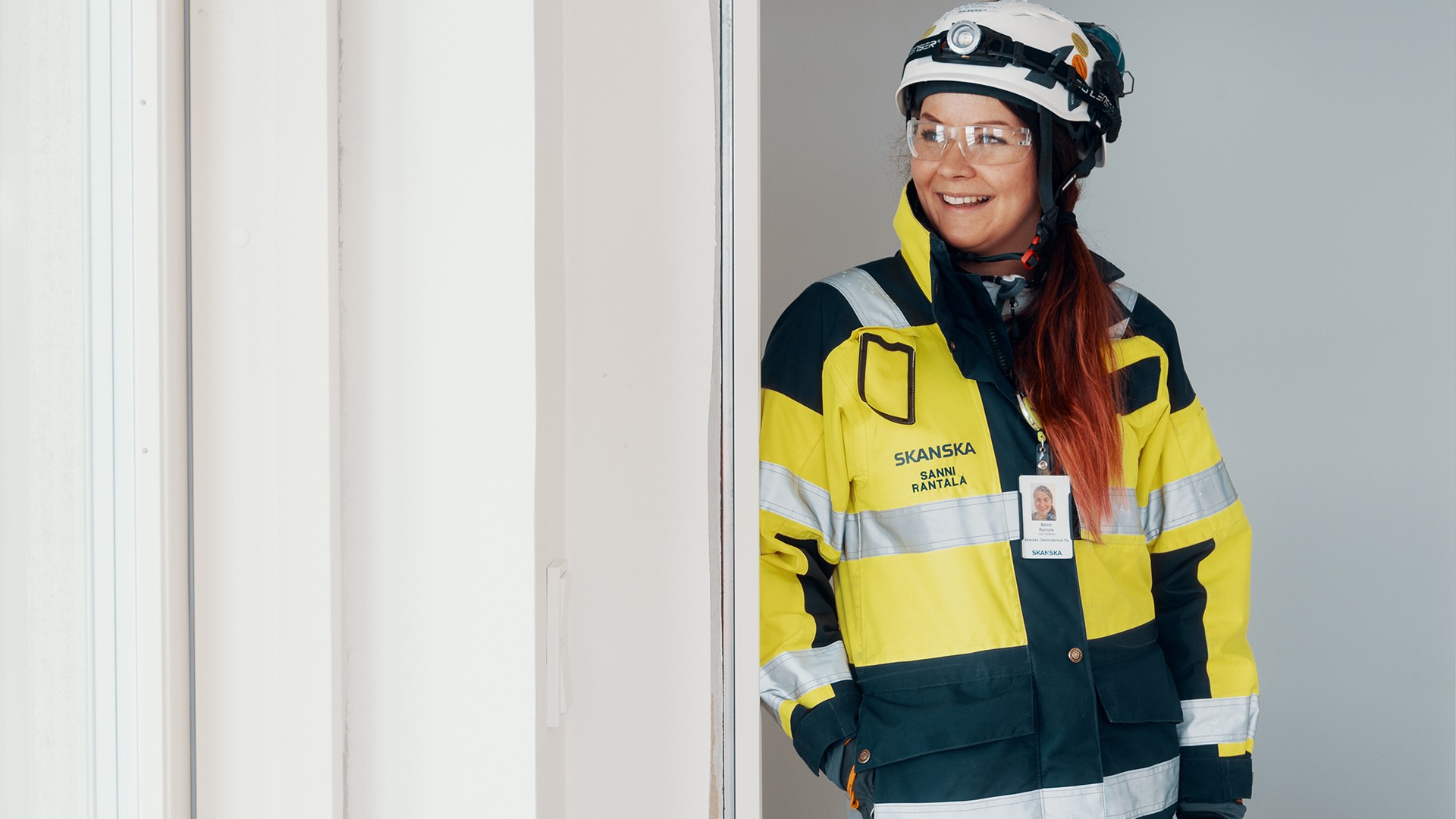 Skanskan työntekijä keltaisessa ja mustassa suojatakissa ja kypärässä seisoo harmaan seinän edessä.