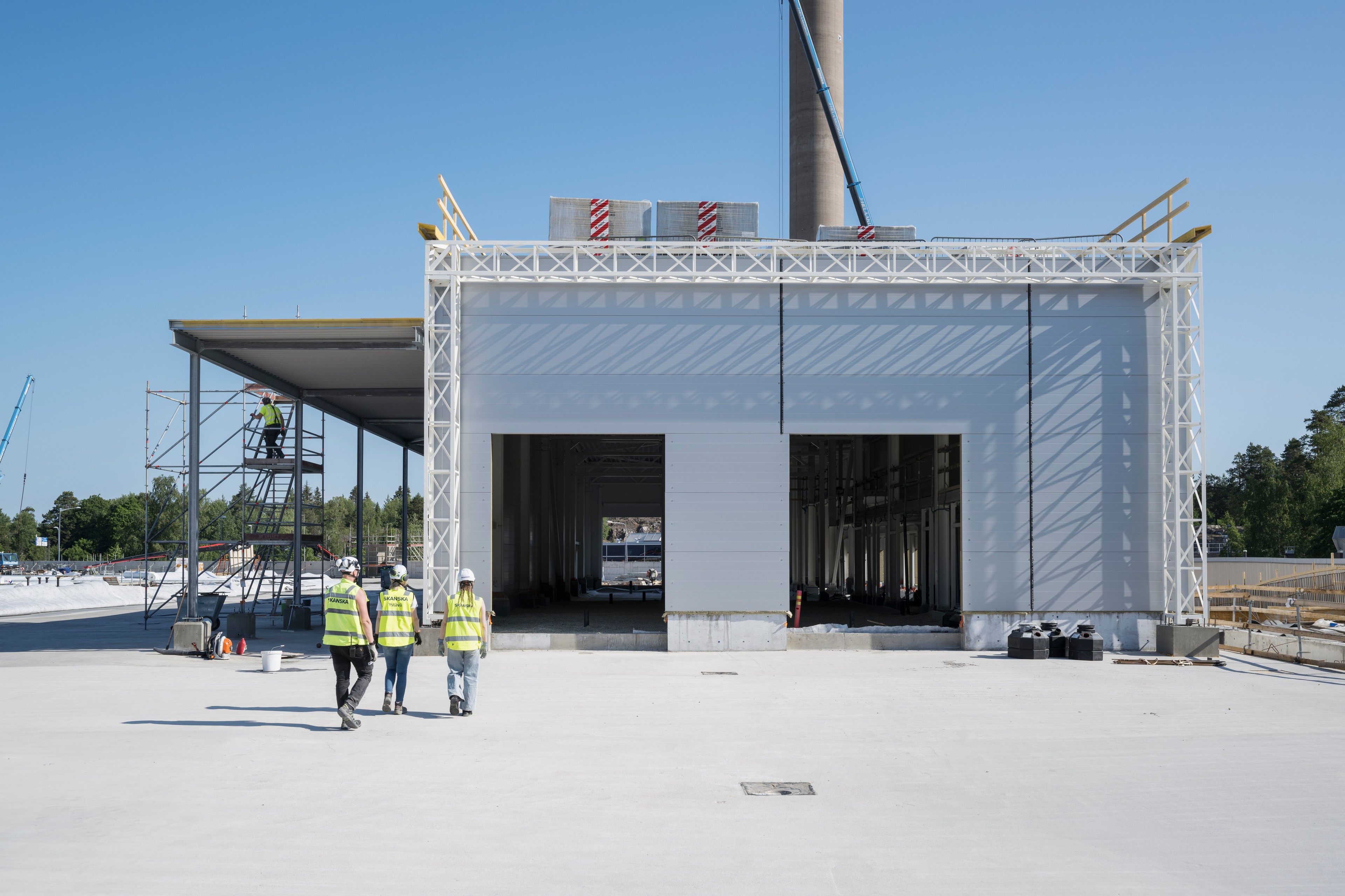 Construction workers in safety gear walk towards a large industrial building under construction.