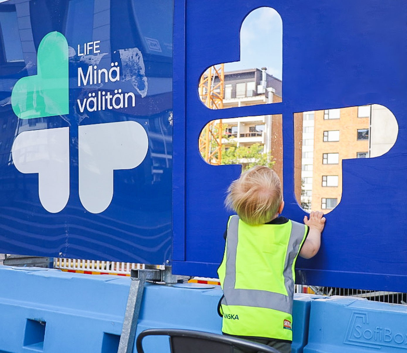 A child in a bright safety vest looks through a blue construction barrier with text LIFE I Care.