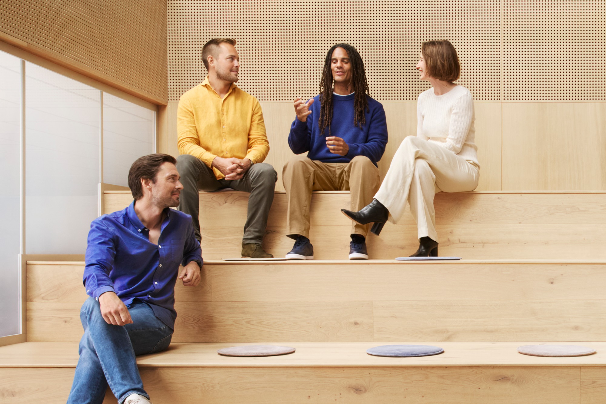 Four people sit on wooden steps, engaged in conversation, wearing casual attire in a modern setting.