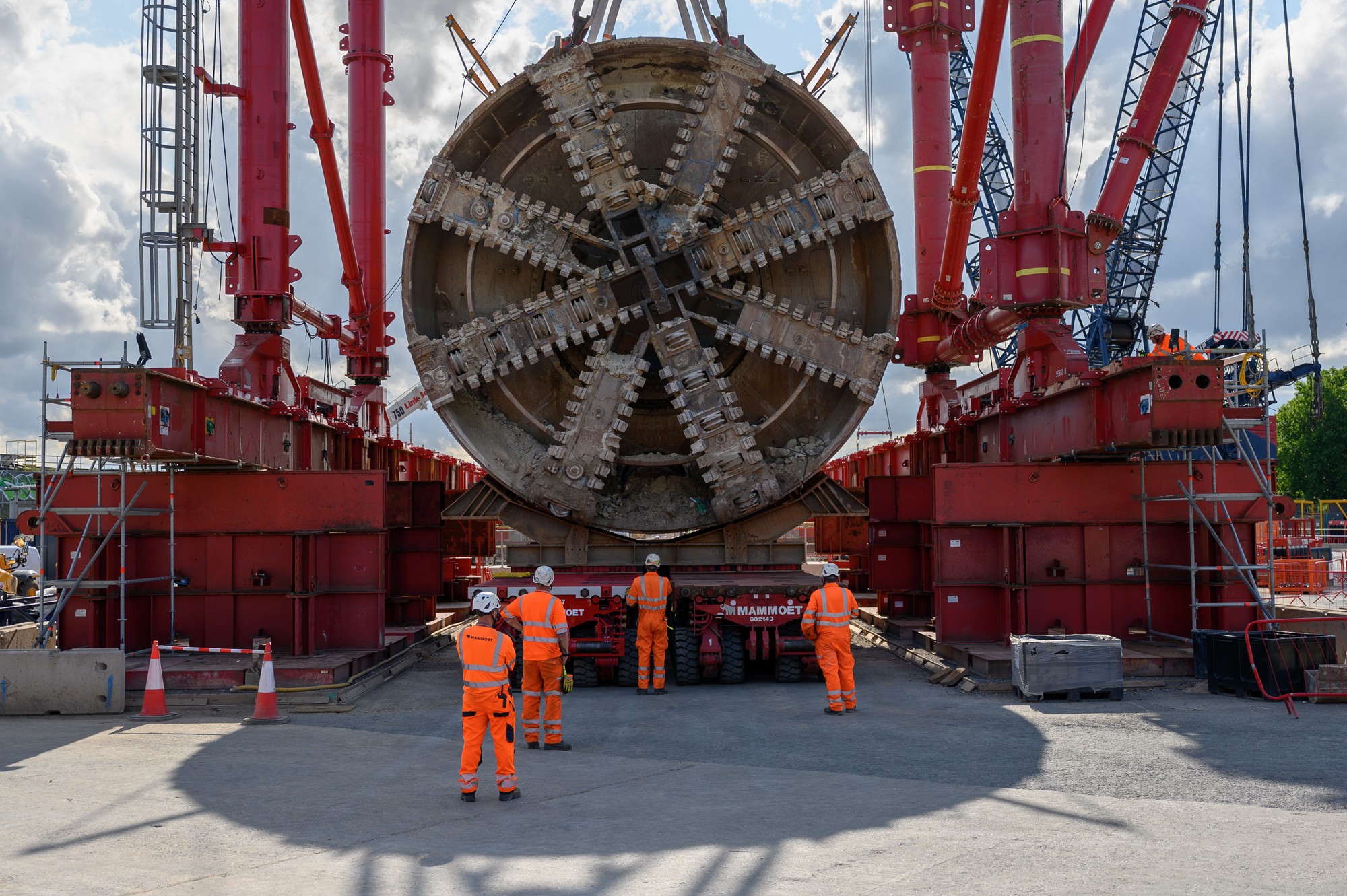 Workers in orange safety gear stand before a large tunnel boring machine at a construction site.