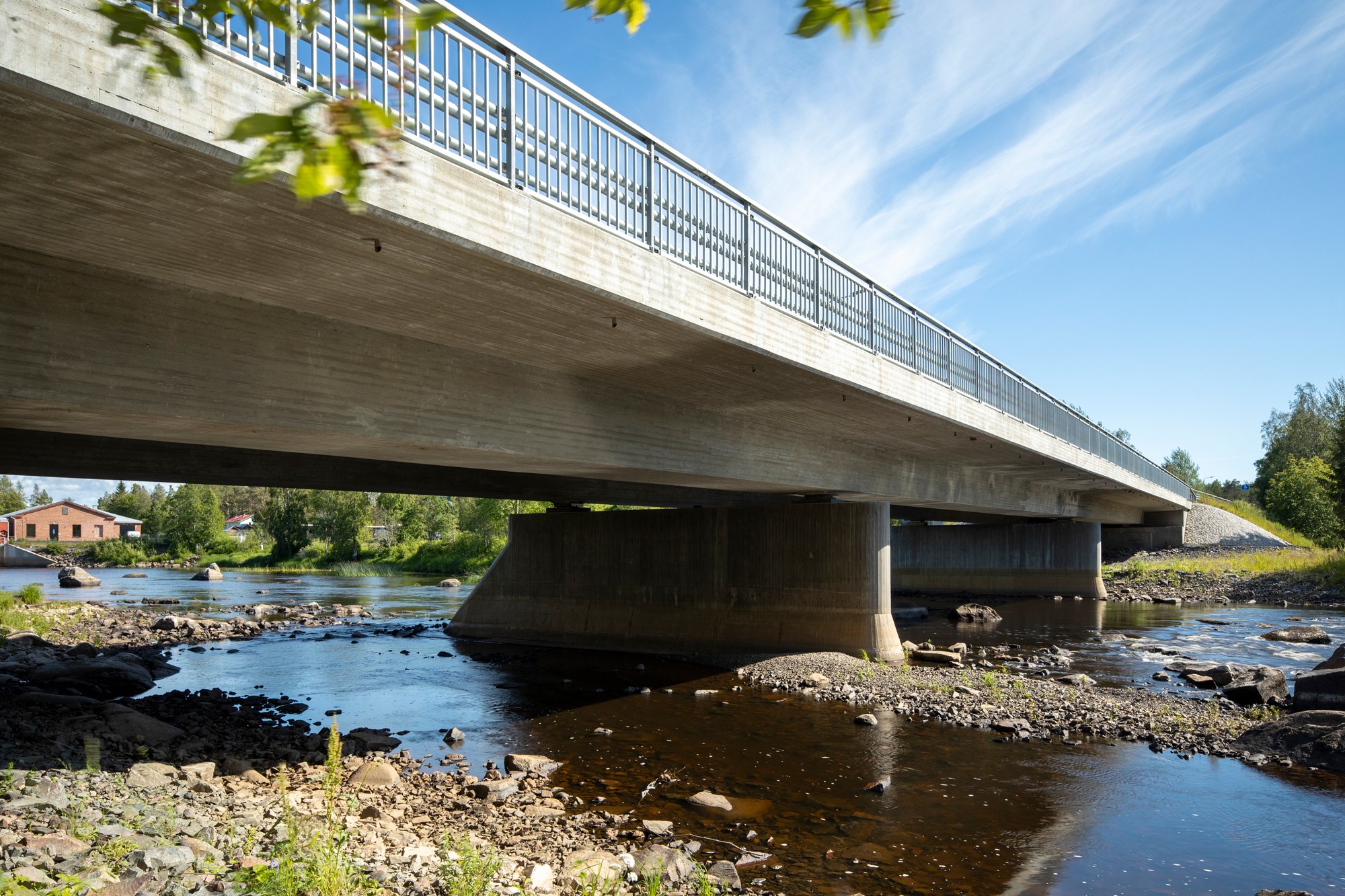 Concrete bridge over a rocky river with blue sky and greenery in the background.