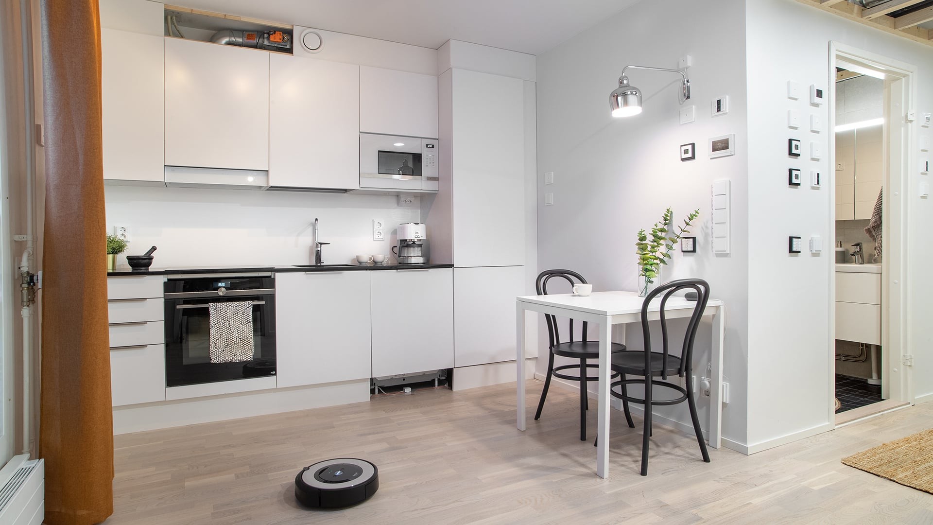 Modern white kitchen with dining table, chairs, and a robotic vacuum on wooden floor.