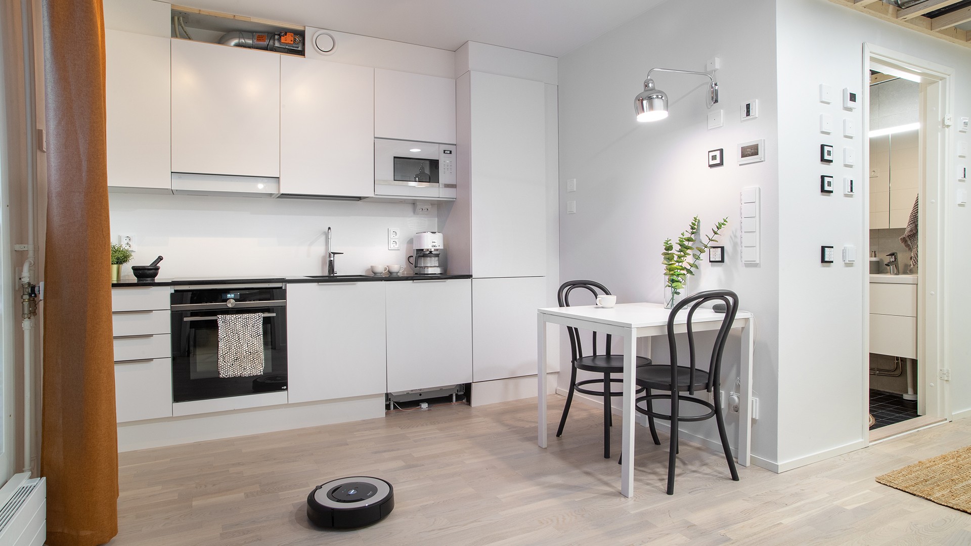 Modern white kitchen with dining table, chairs, and a robotic vacuum on wooden floor.