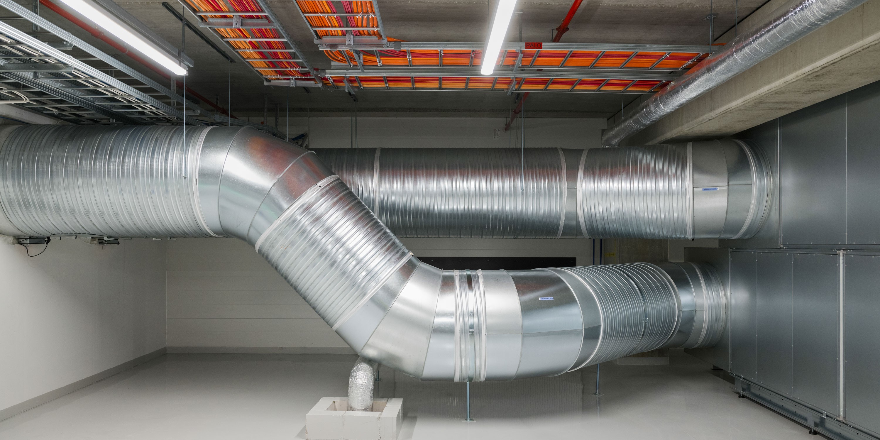 Industrial HVAC duct system in a commercial building with metallic pipes and overhead lighting.